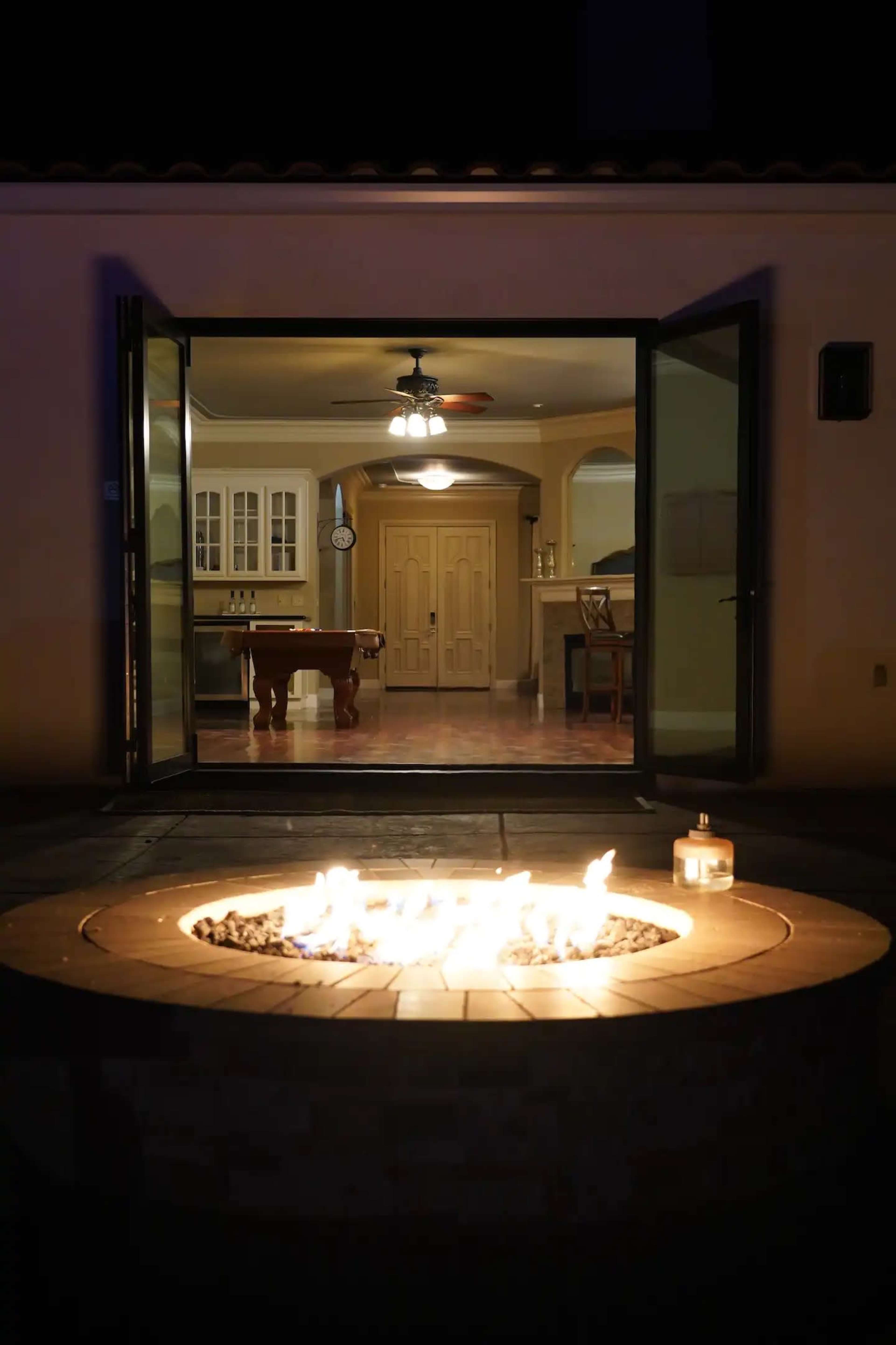 A stone fire pit with flames is in the foreground, while large open doors reveal a well-lit kitchen and dining area in the background.