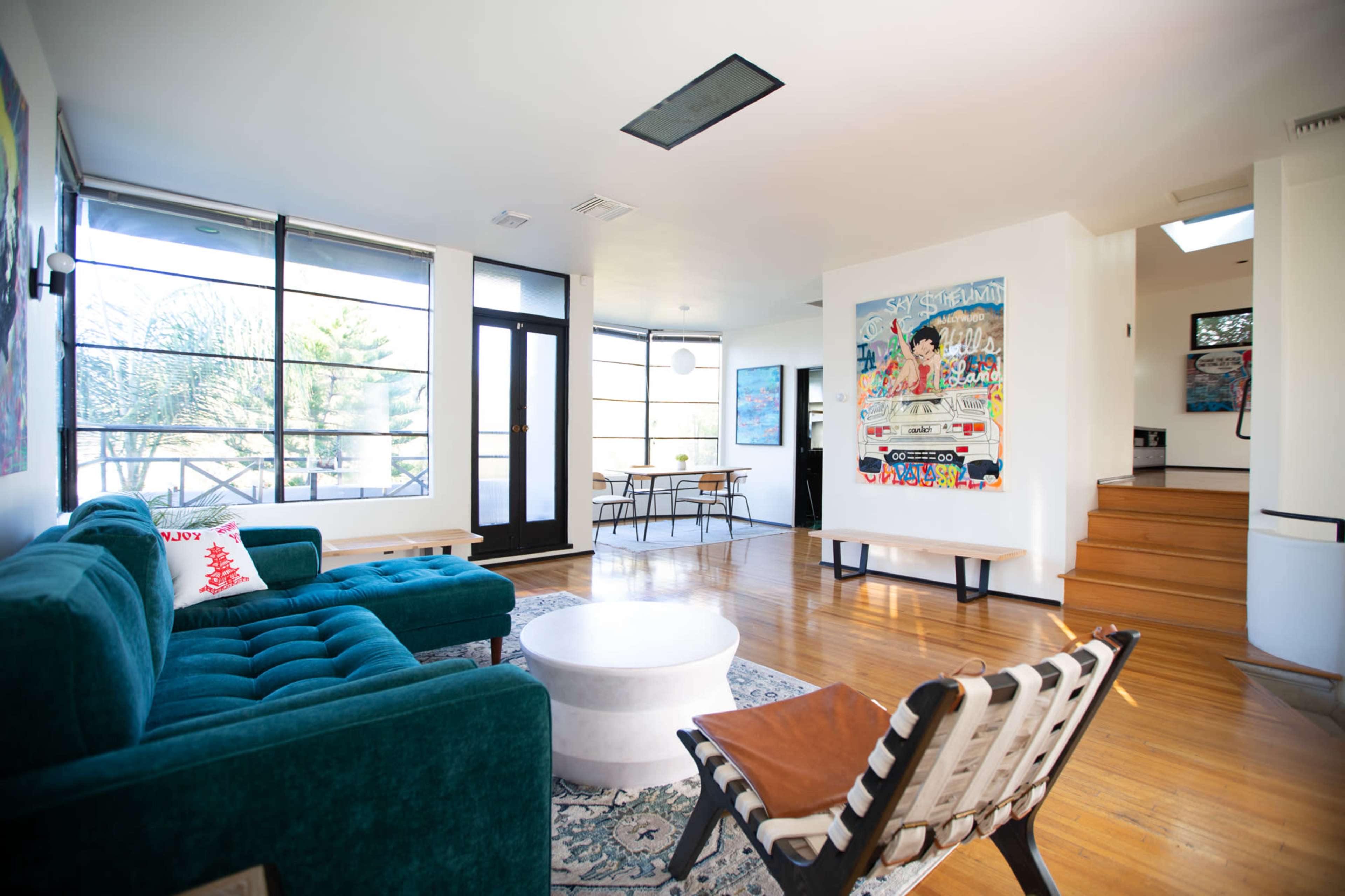 The image shows a modern living room with a teal sofa, a round coffee table, and large windows allowing natural light, leading to a dining area and a staircase.