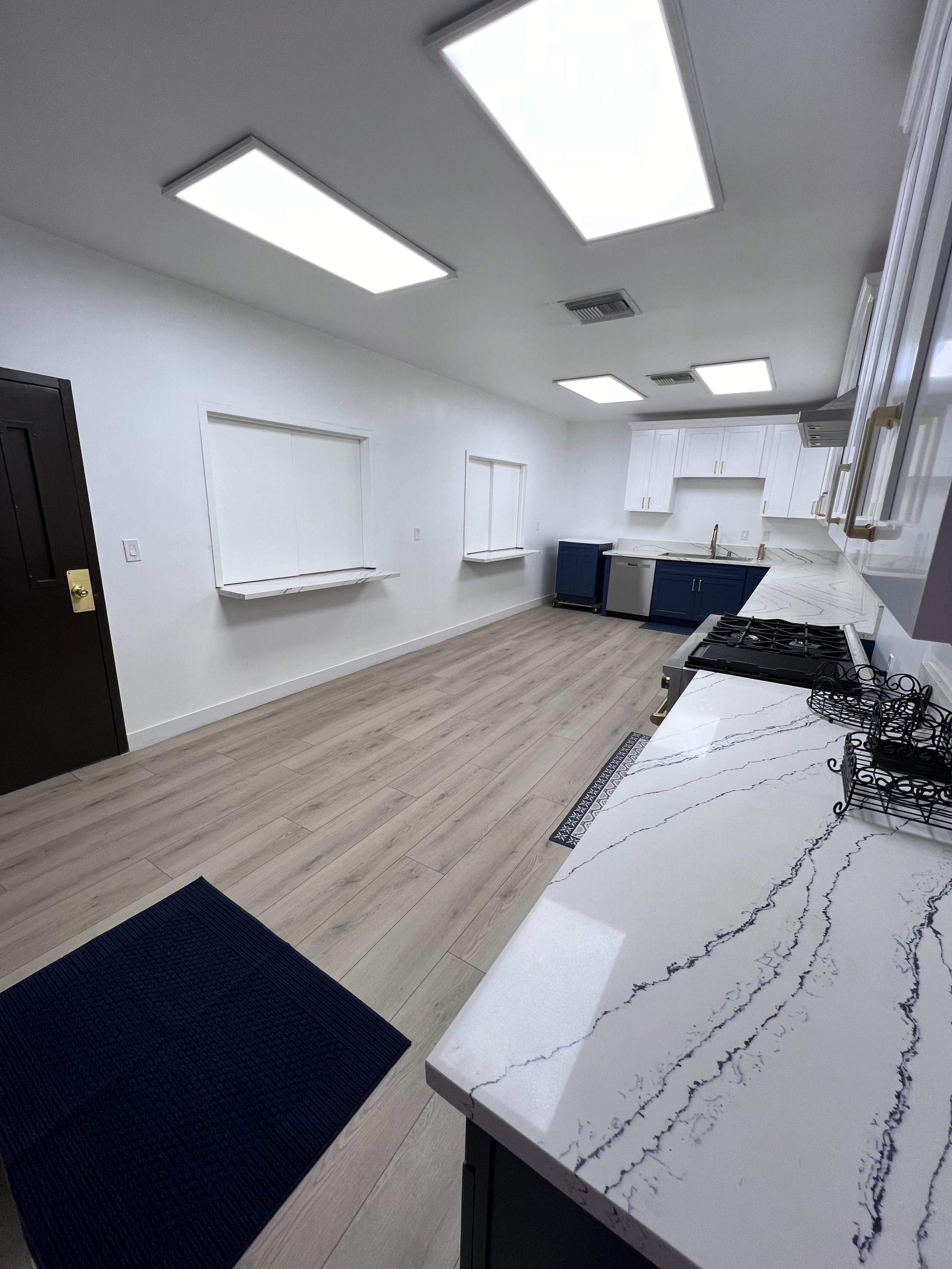 The image shows a spacious kitchen with white walls, blue cabinetry, and a marble countertop, featuring skylights and an empty floor space.
