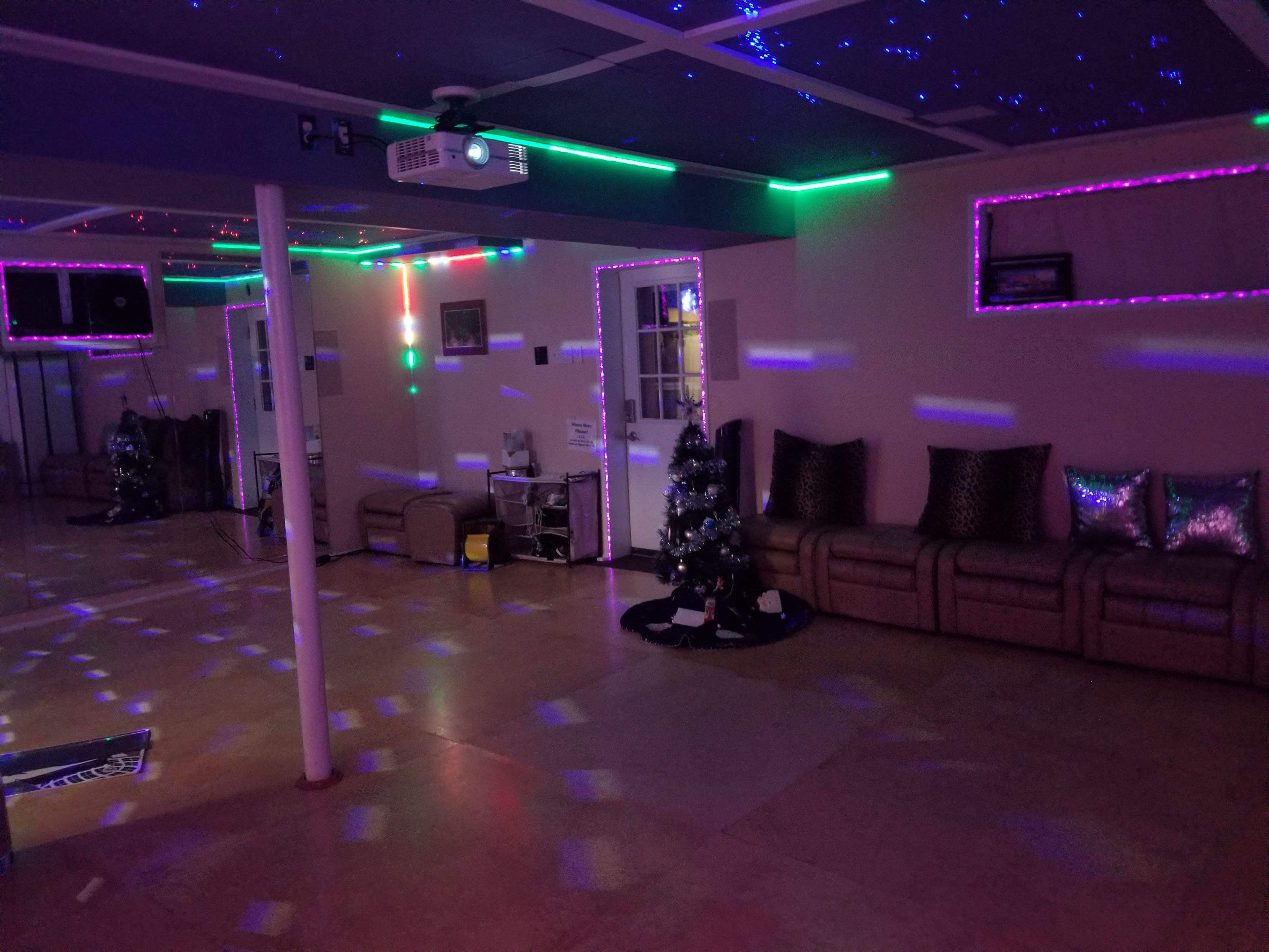 A spacious room with colorful lights, a projector on the ceiling, a Christmas tree in one corner, and a seating area with metallic pillows.