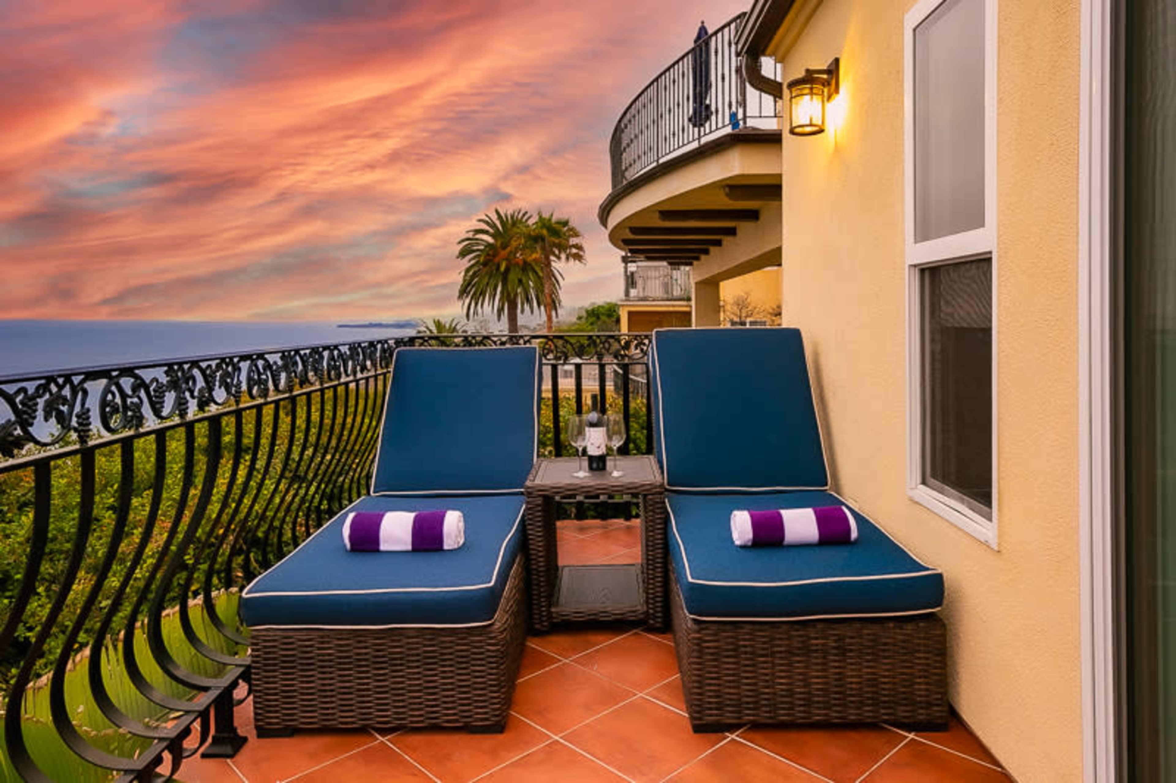 A balcony with two blue lounge chairs and a small table, overlooking a scenic view of the ocean and palm trees under a colorful sky.