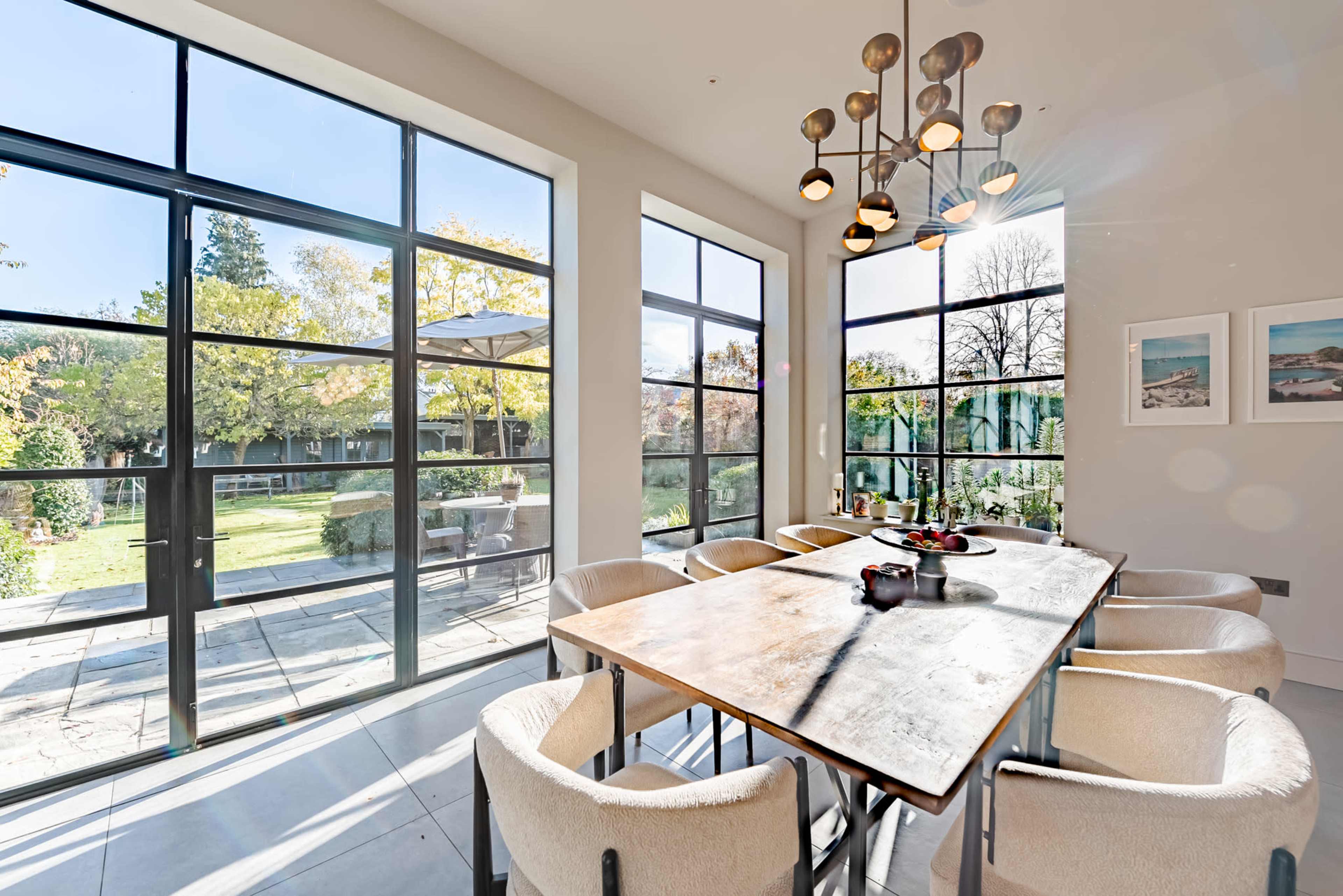 A modern dining area features a large wooden table surrounded by upholstered chairs, with large windows offering views of an outdoor garden.