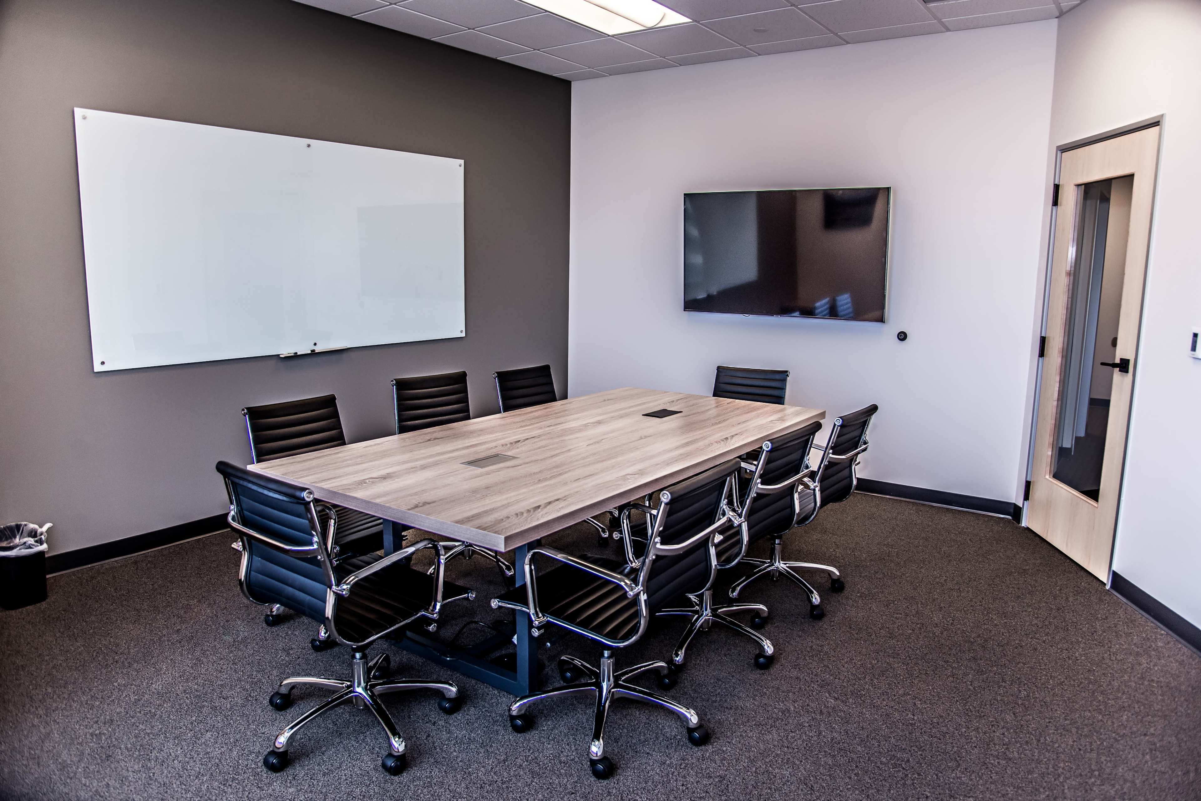 A modern conference room features a large rectangular table surrounded by black chairs, with a whiteboard and a television mounted on the wall.