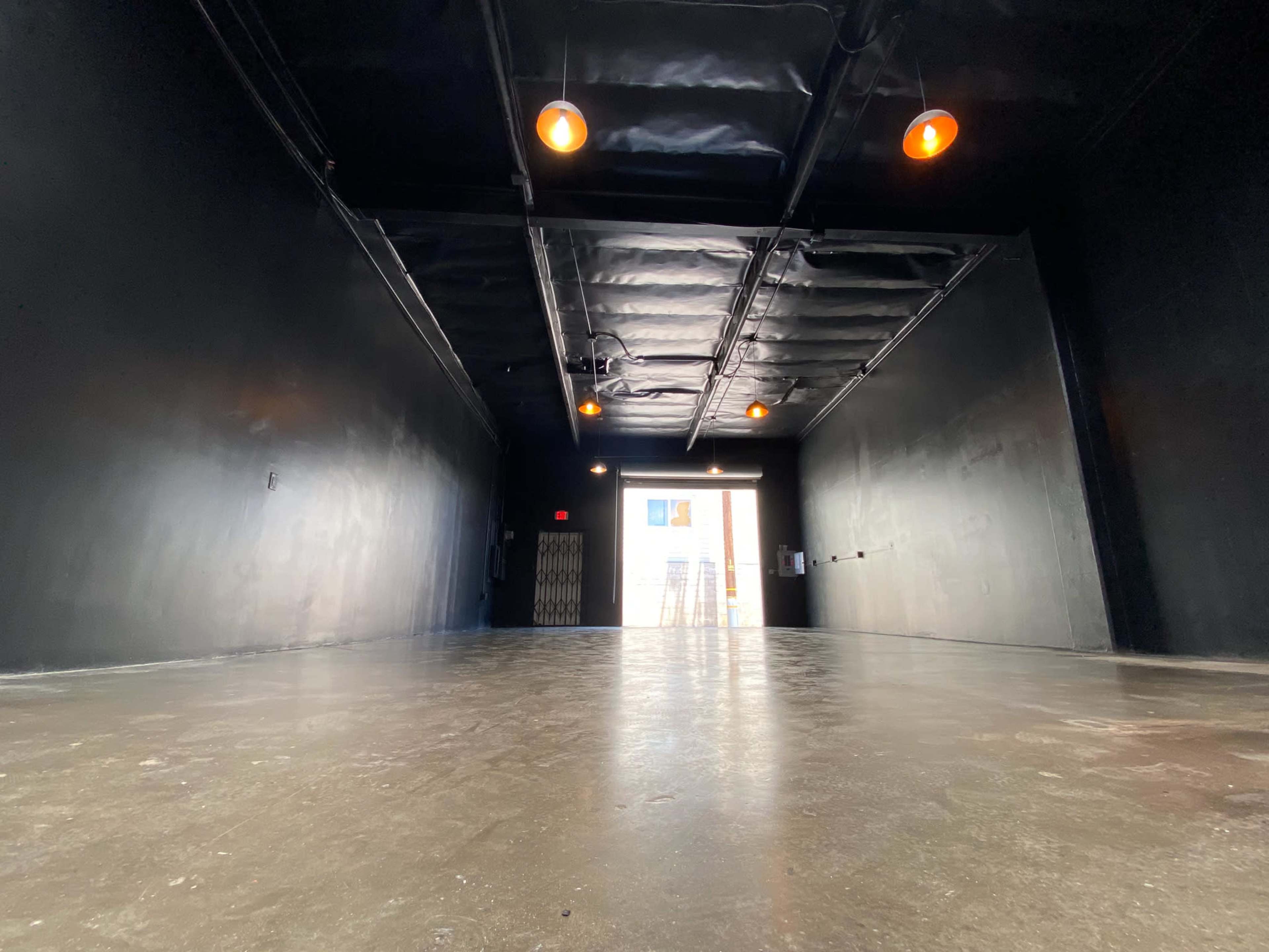 The image shows an empty, dimly lit hallway with a black ceiling, walls, and a concrete floor, leading to a door that opens to an outdoor area.