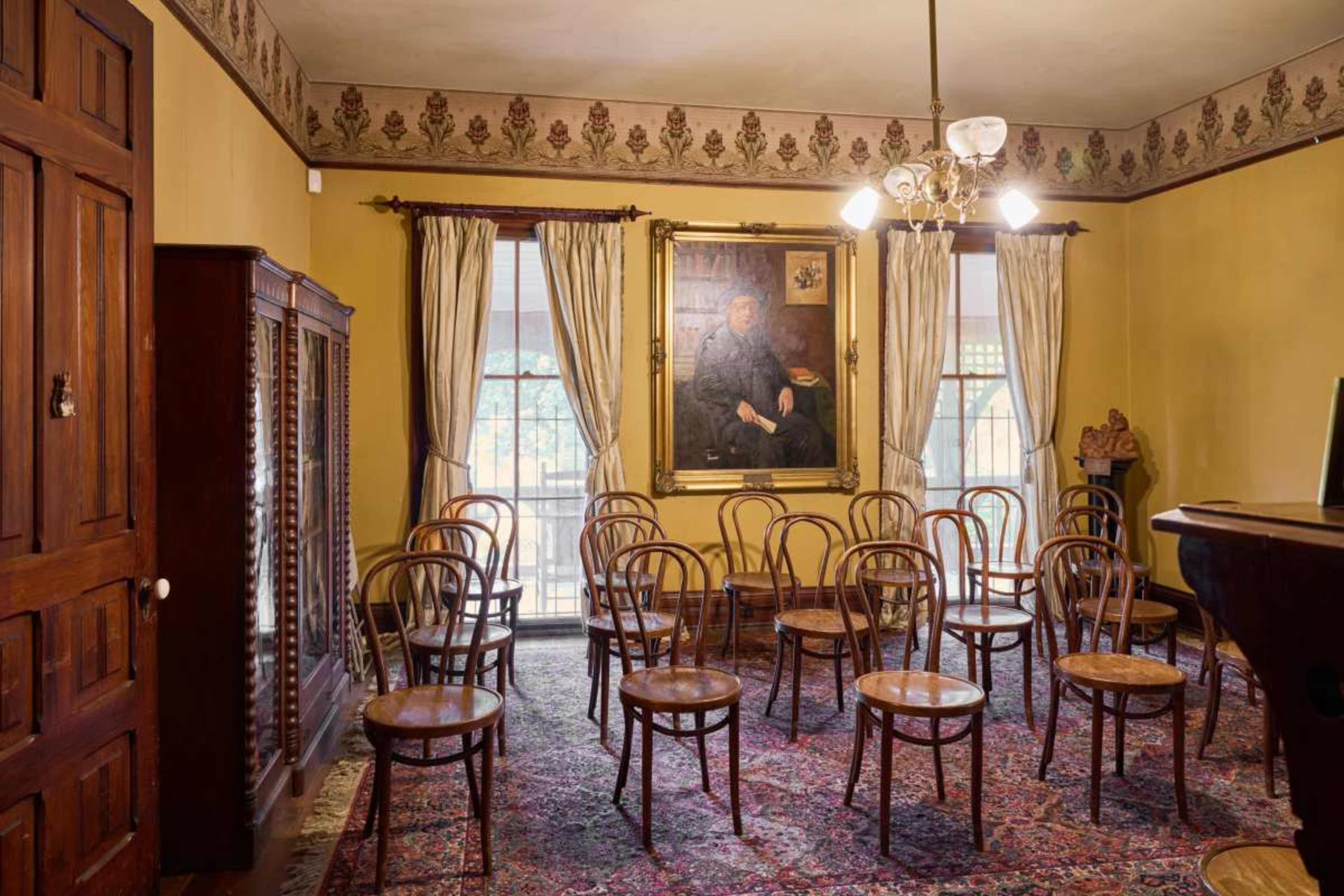 The room features a collection of wooden chairs arranged in a circle around a large portrait, with yellow walls and decorative trim.