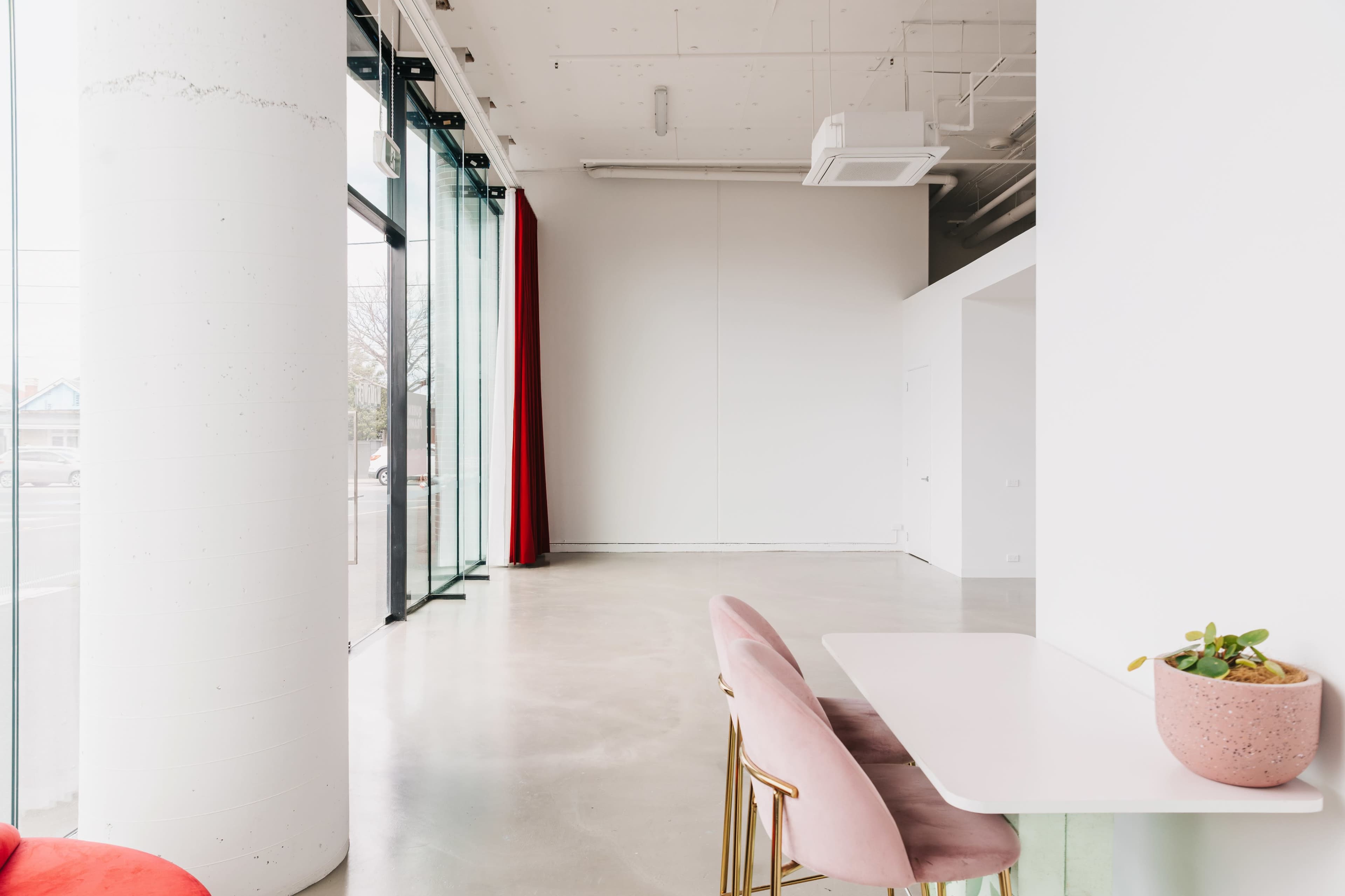 The image shows a modern, minimalist interior featuring a pink chair and a small table near large glass windows with red curtains.