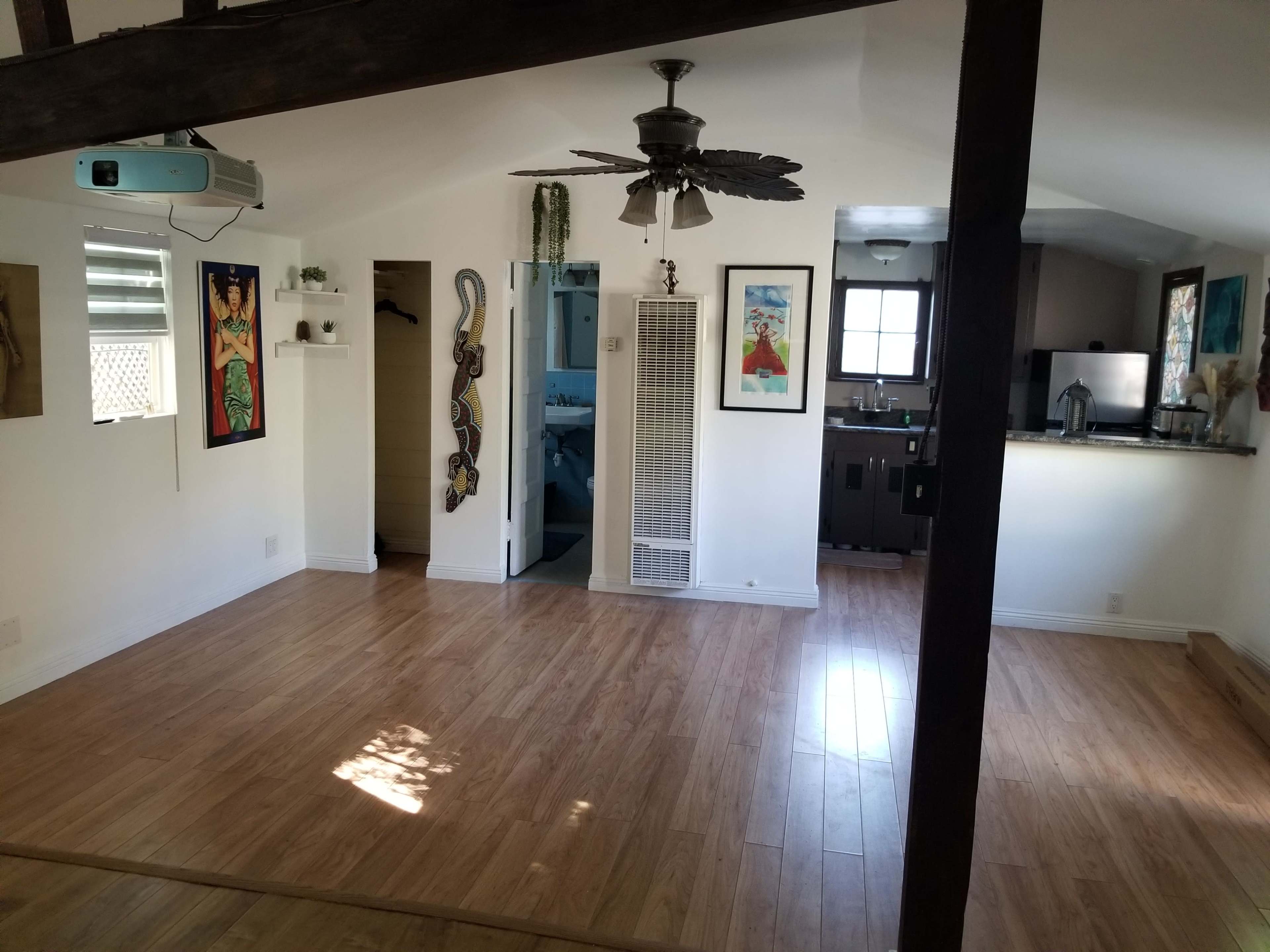 The image shows a spacious room with wooden flooring, featuring a ceiling fan, a wall-mounted projector, and doorways leading to other areas.