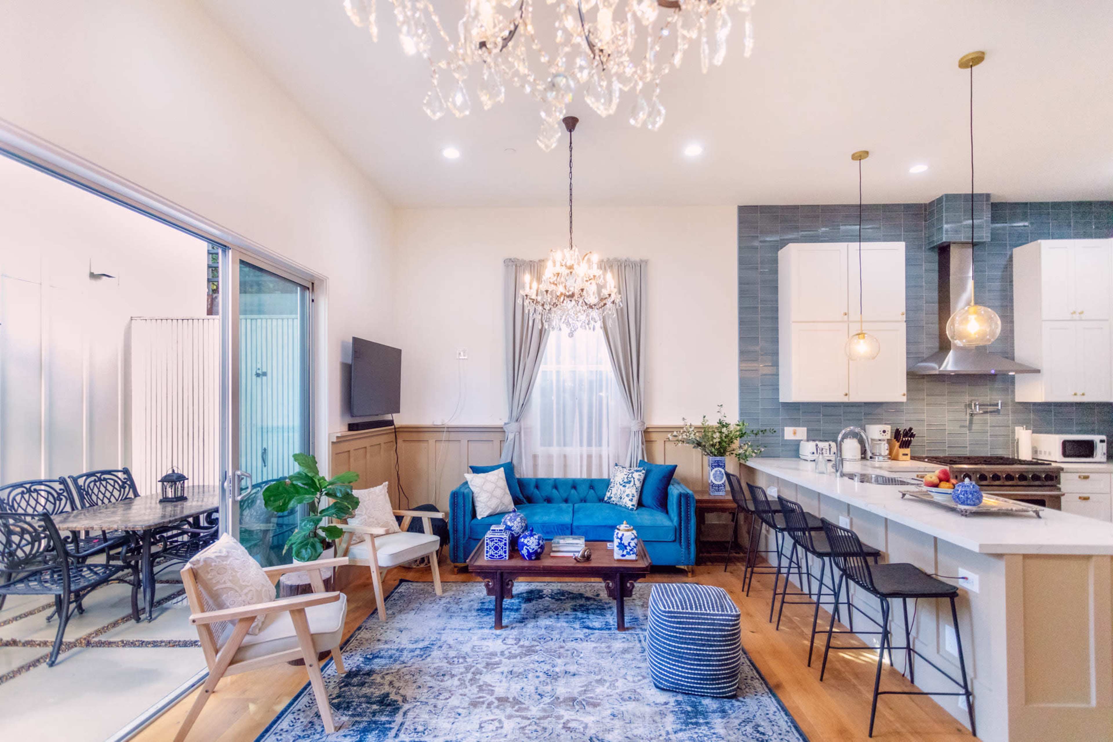 A bright living area features a blue sofa, a coffee table, and a kitchen with modern appliances, all illuminated by a chandelier and pendant lights.
