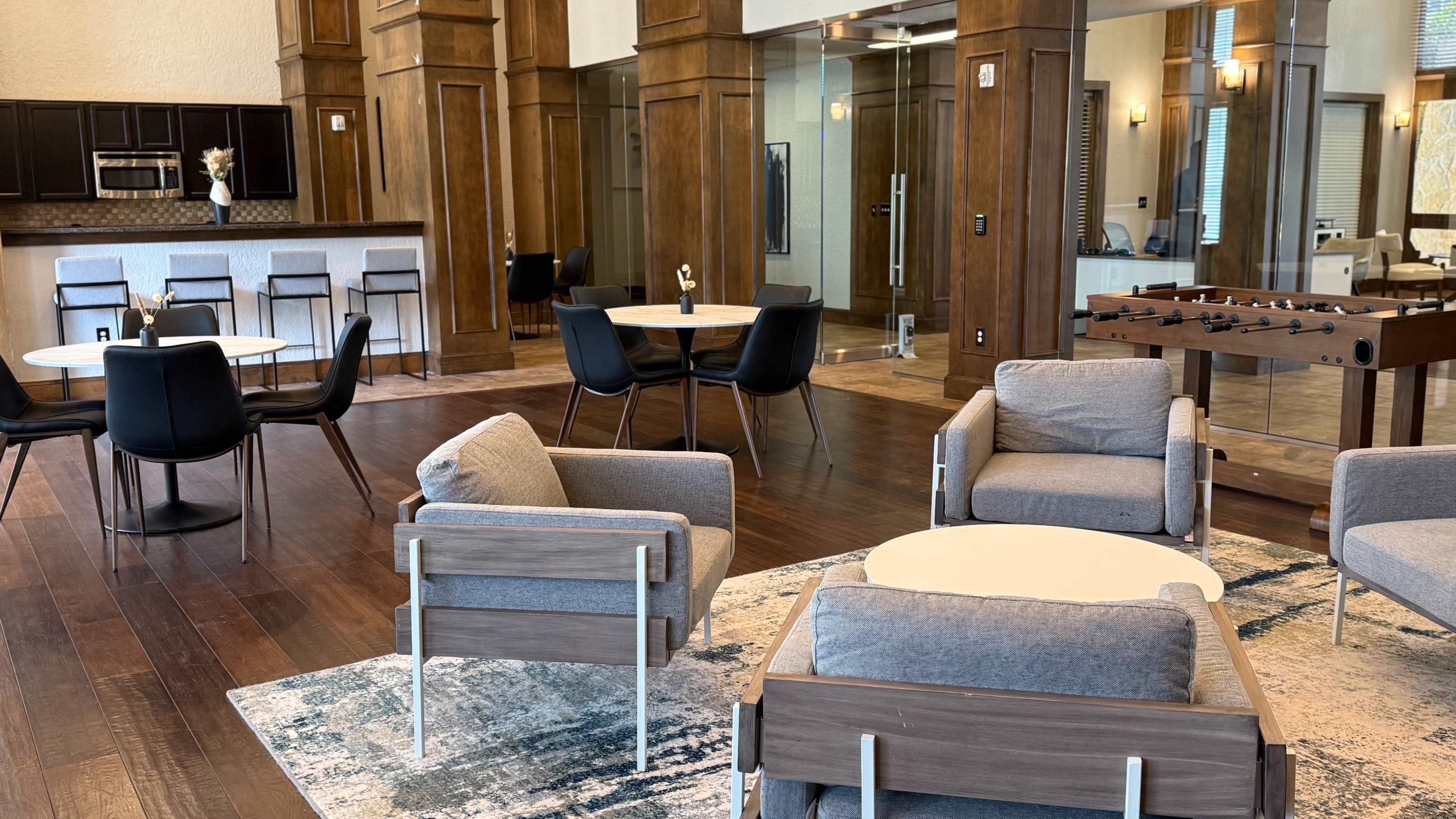 The image depicts a stylish communal area featuring various seating arrangements, including gray chairs and tables, with a foosball table in the corner and a large window allowing natural light.