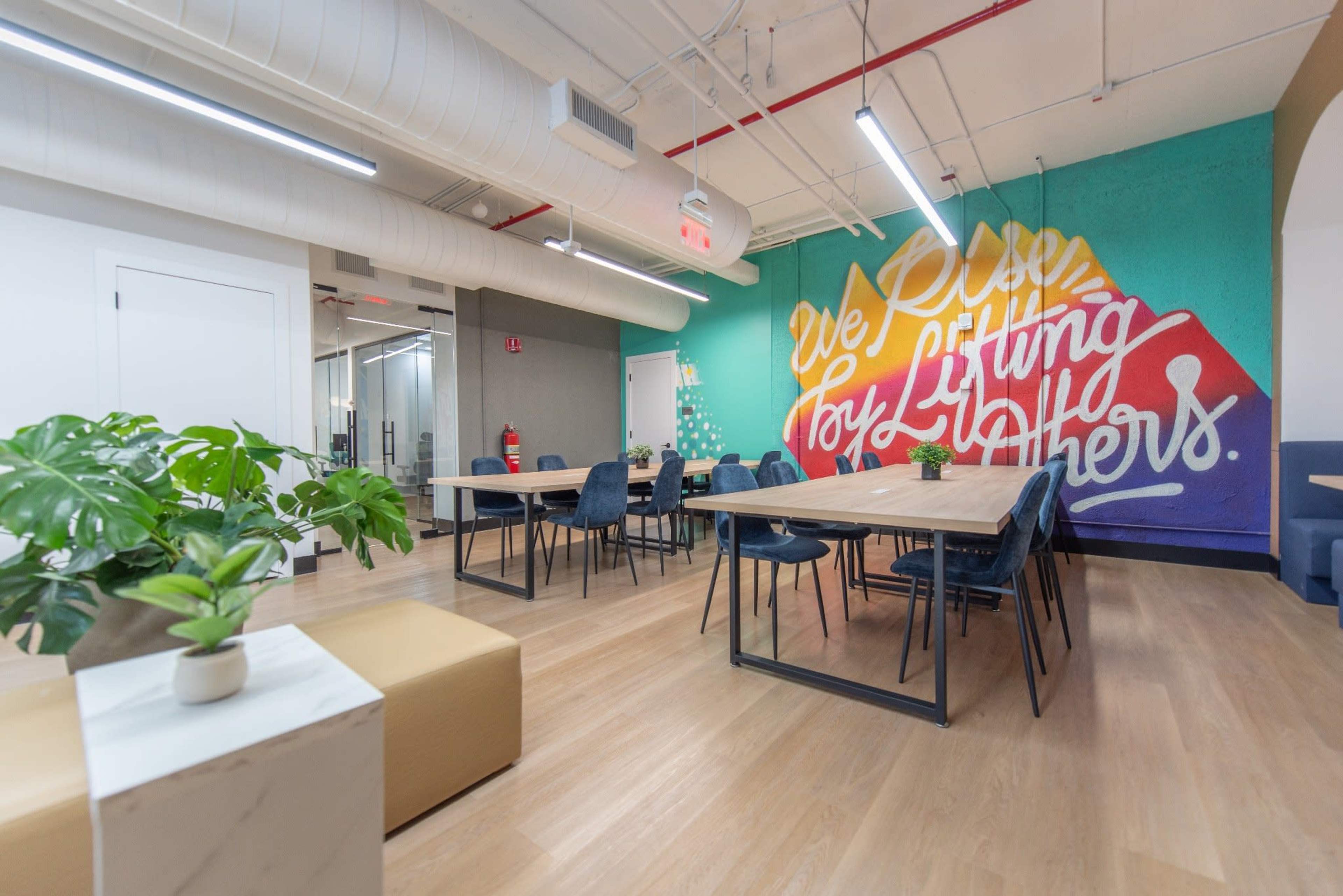 The image shows a modern, open office space with several wooden tables and chairs, featuring a colorful mural on one wall that reads “We Live for Lifting Others.”
