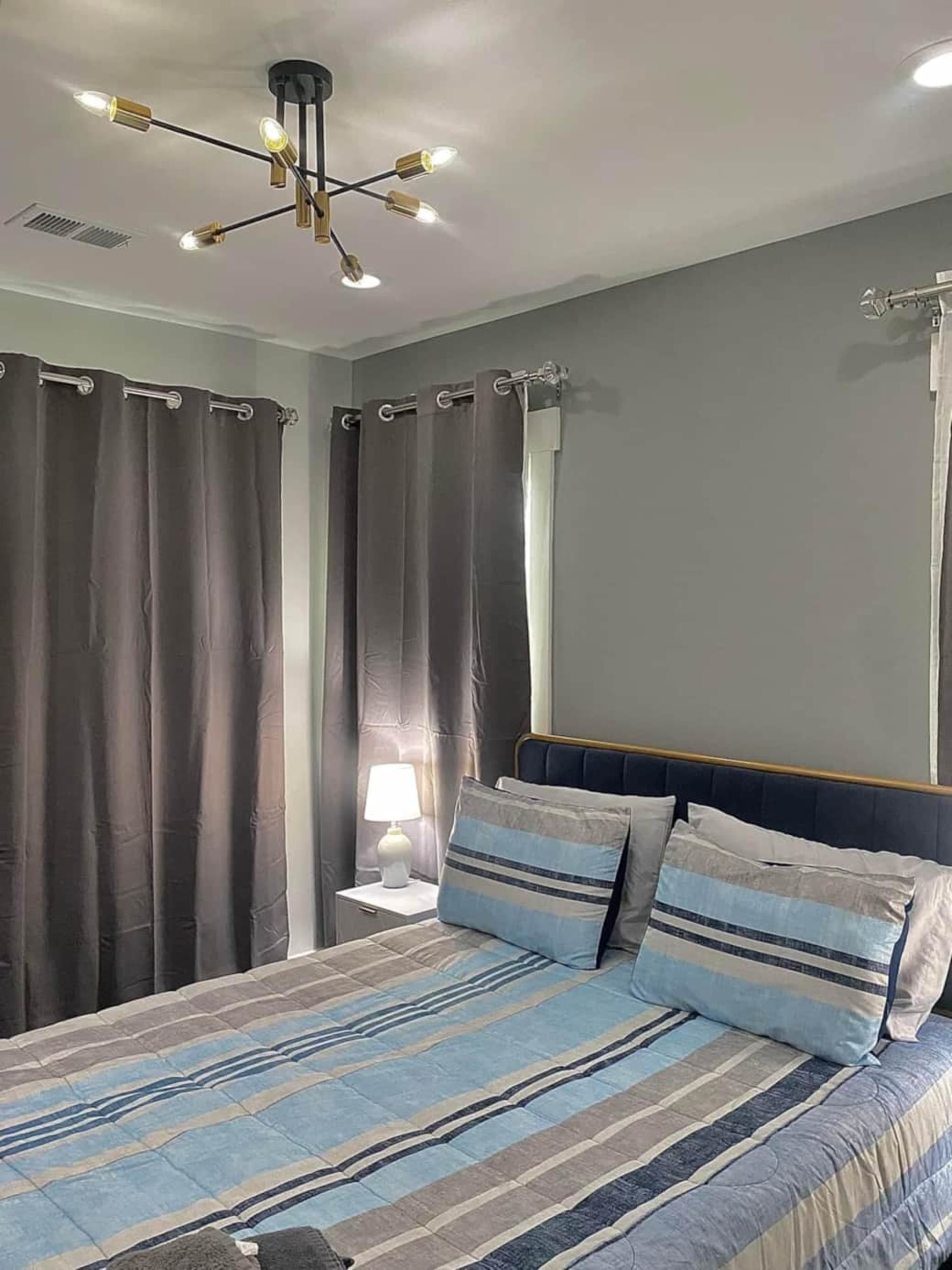 A bedroom with a bed featuring blue and gray striped bedding, two dark curtains covering the windows, and a small bedside lamp on a white nightstand.
