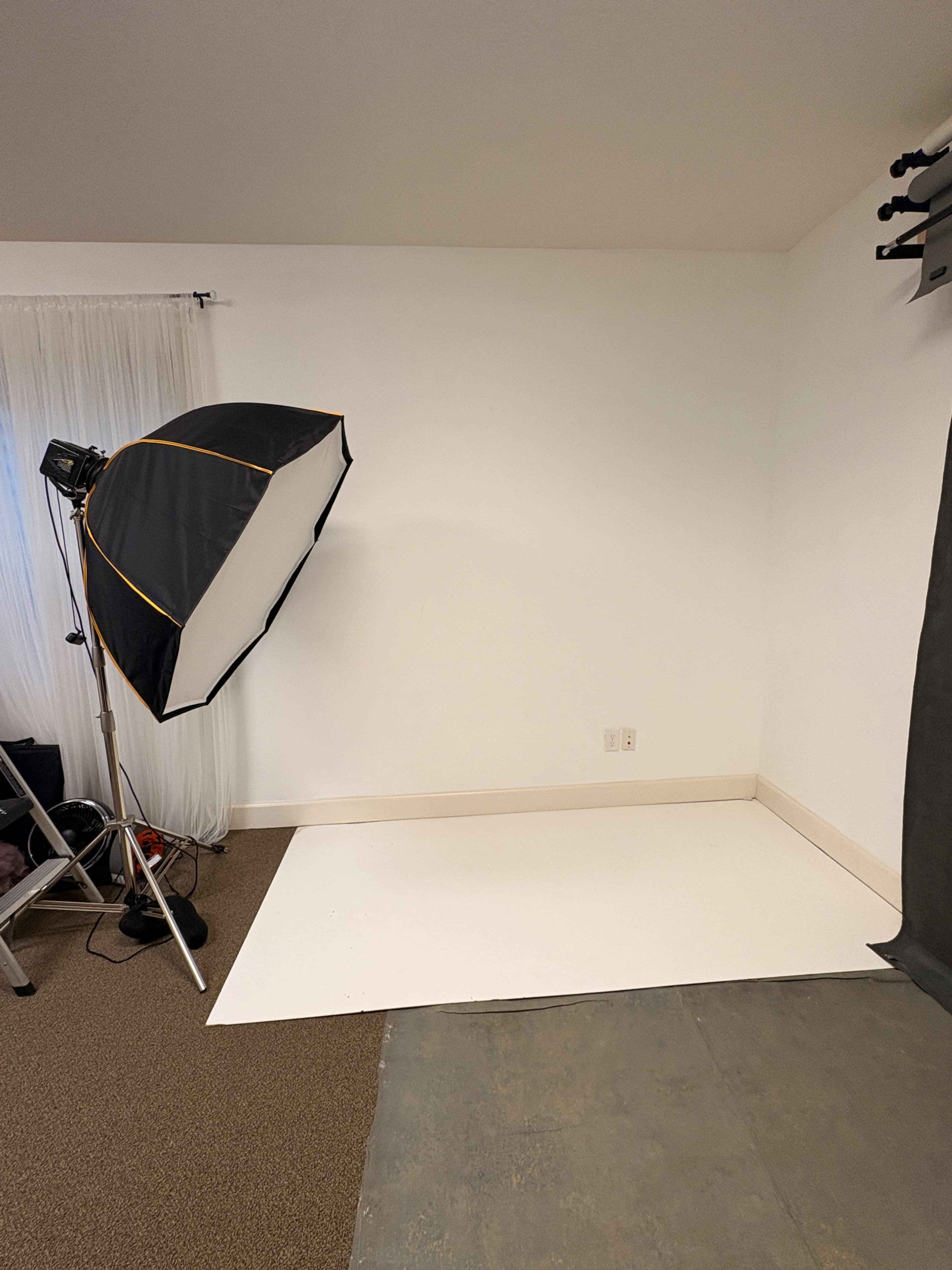 The image shows a photography studio setup with a large softbox light on a stand and a plain white backdrop positioned against a wall.