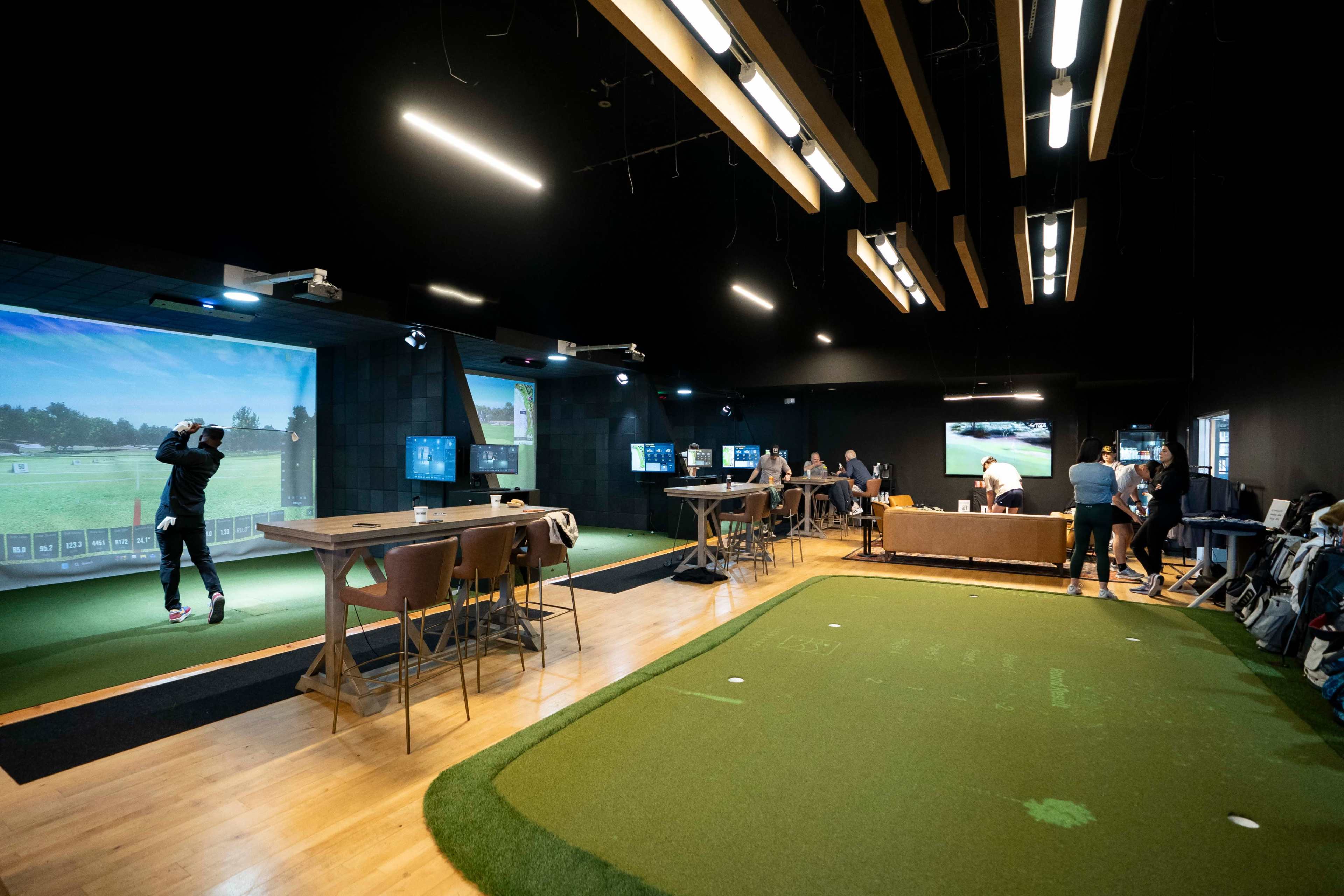 The image shows an indoor golf simulation facility with multiple bays, various patrons practicing their swings, and a putting area on a green carpet.