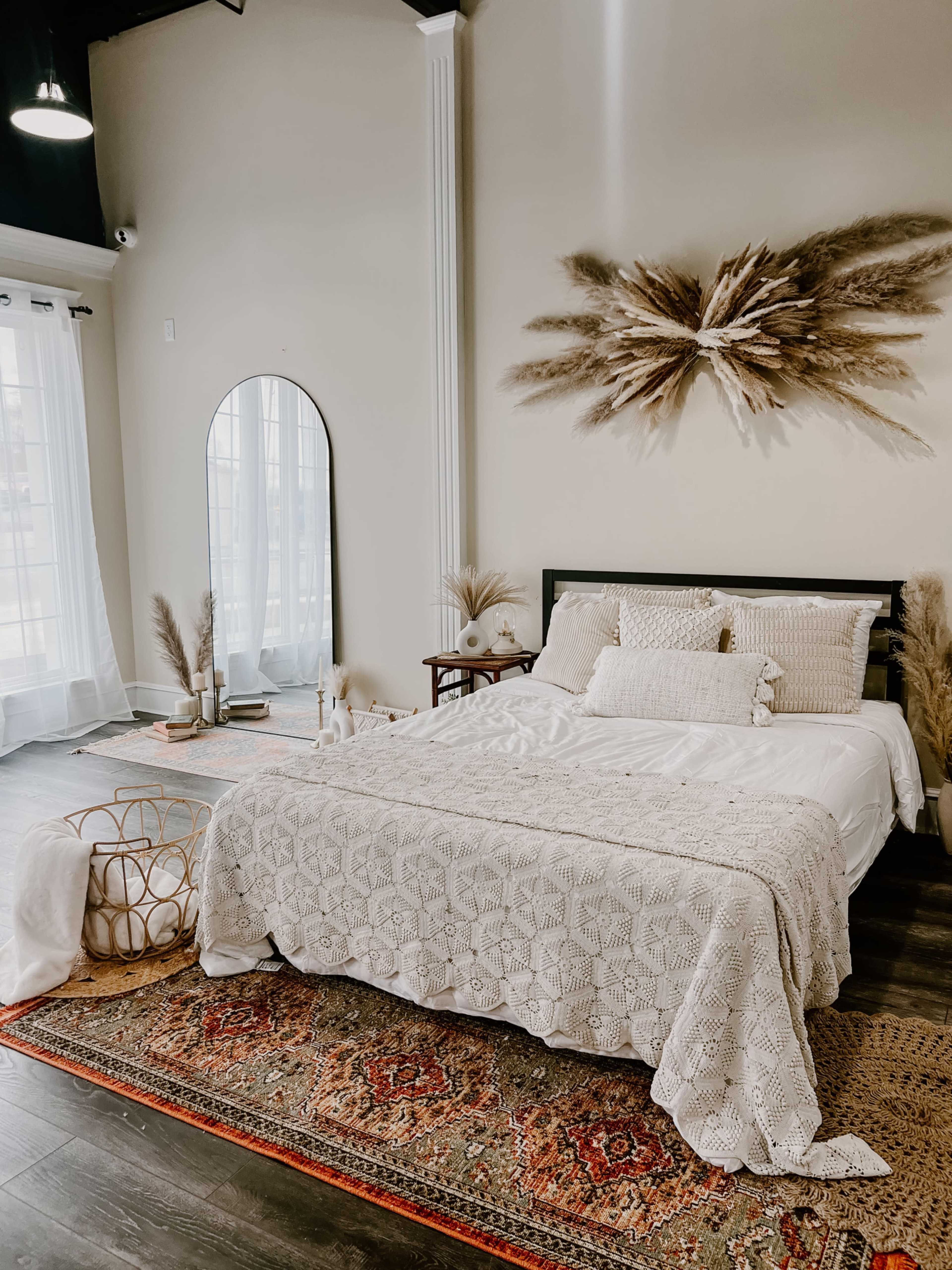 A neatly arranged bedroom features a central bed with a textured white blanket, decorative pillows, and plants, alongside a large mirror and an ornate rug.