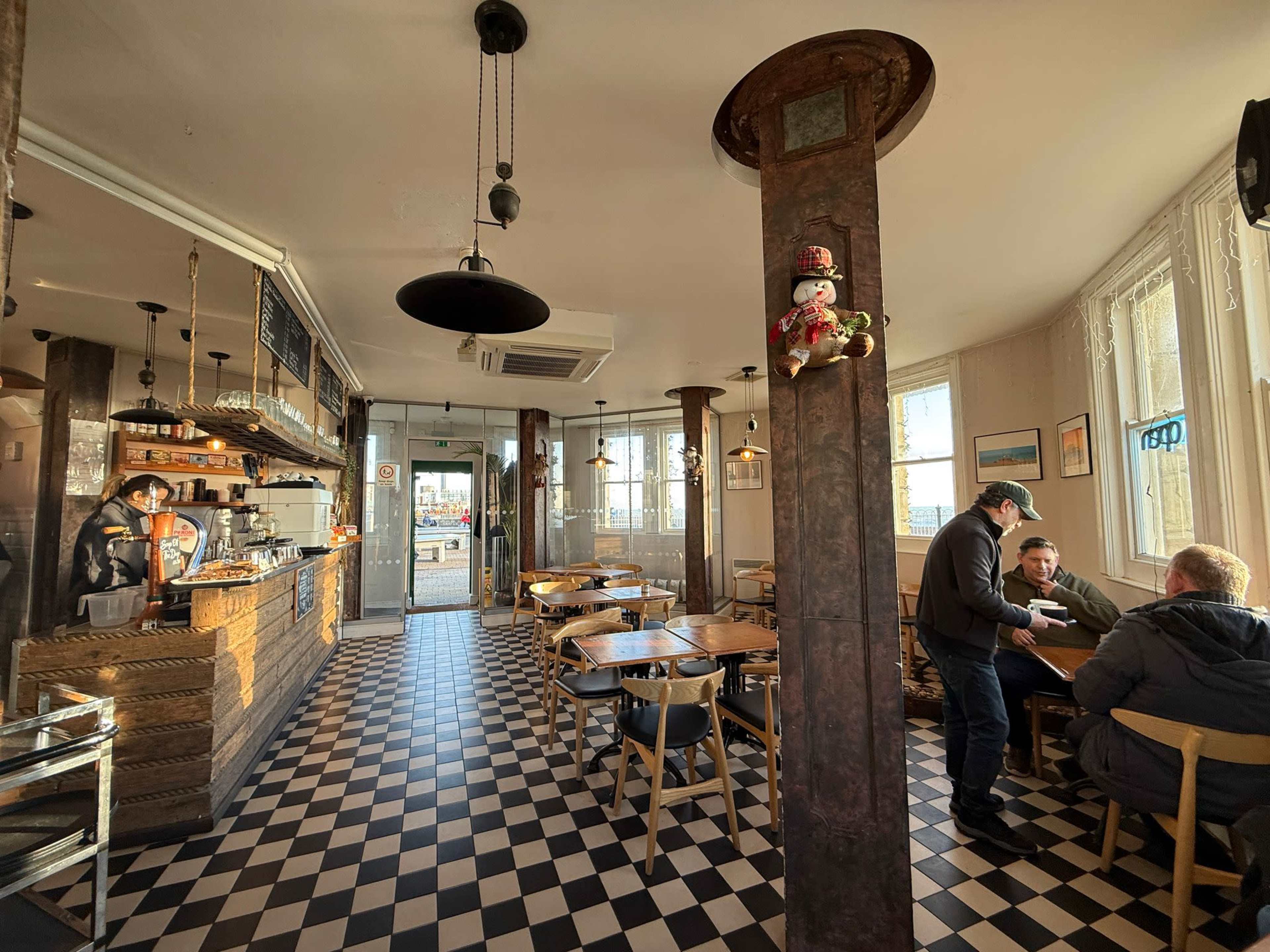 The image shows the interior of a café with a checkered floor, wooden tables, and a counter where a barista is serving customers.