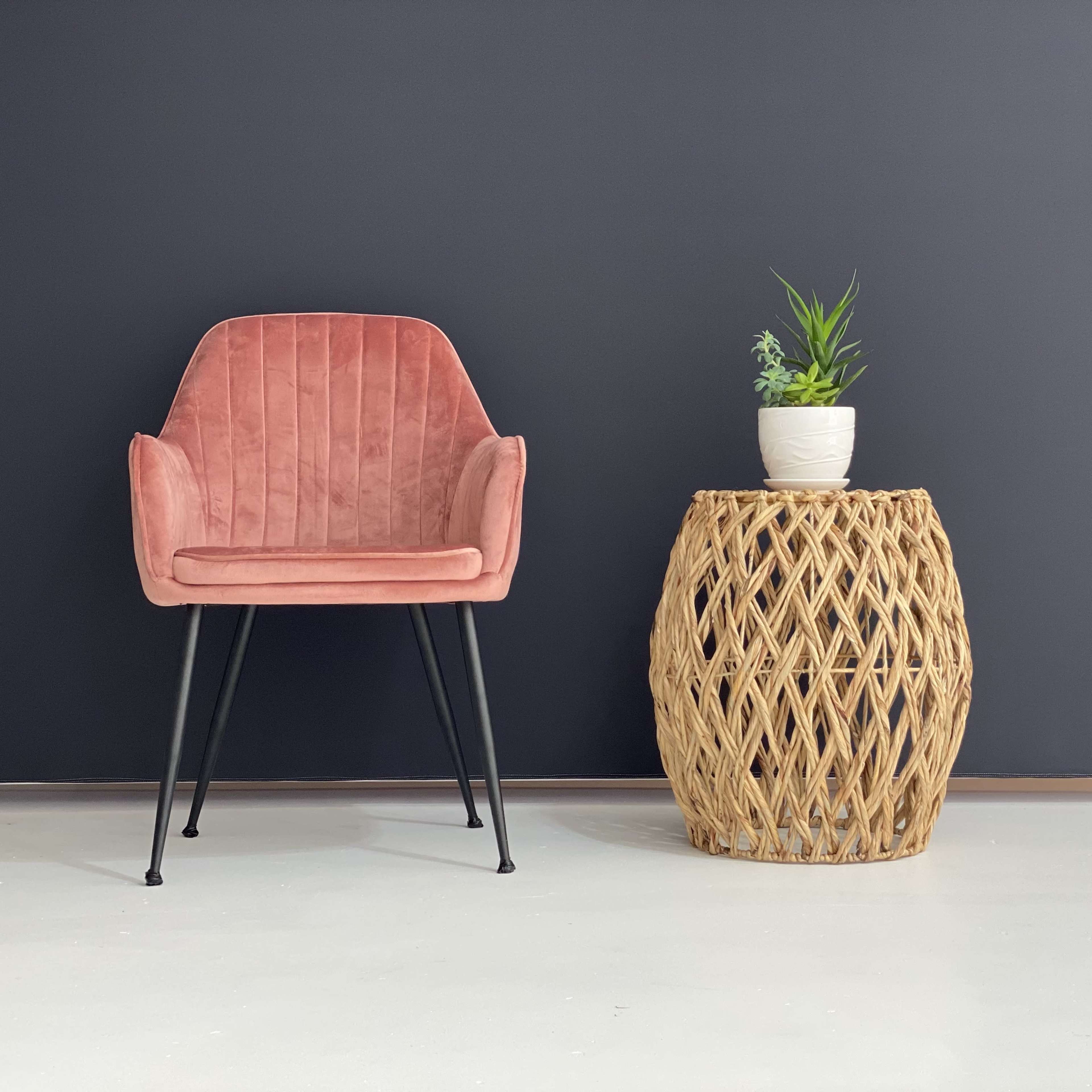 A pink velvet chair is positioned next to a woven wicker side table topped with a potted plant against a dark wall.