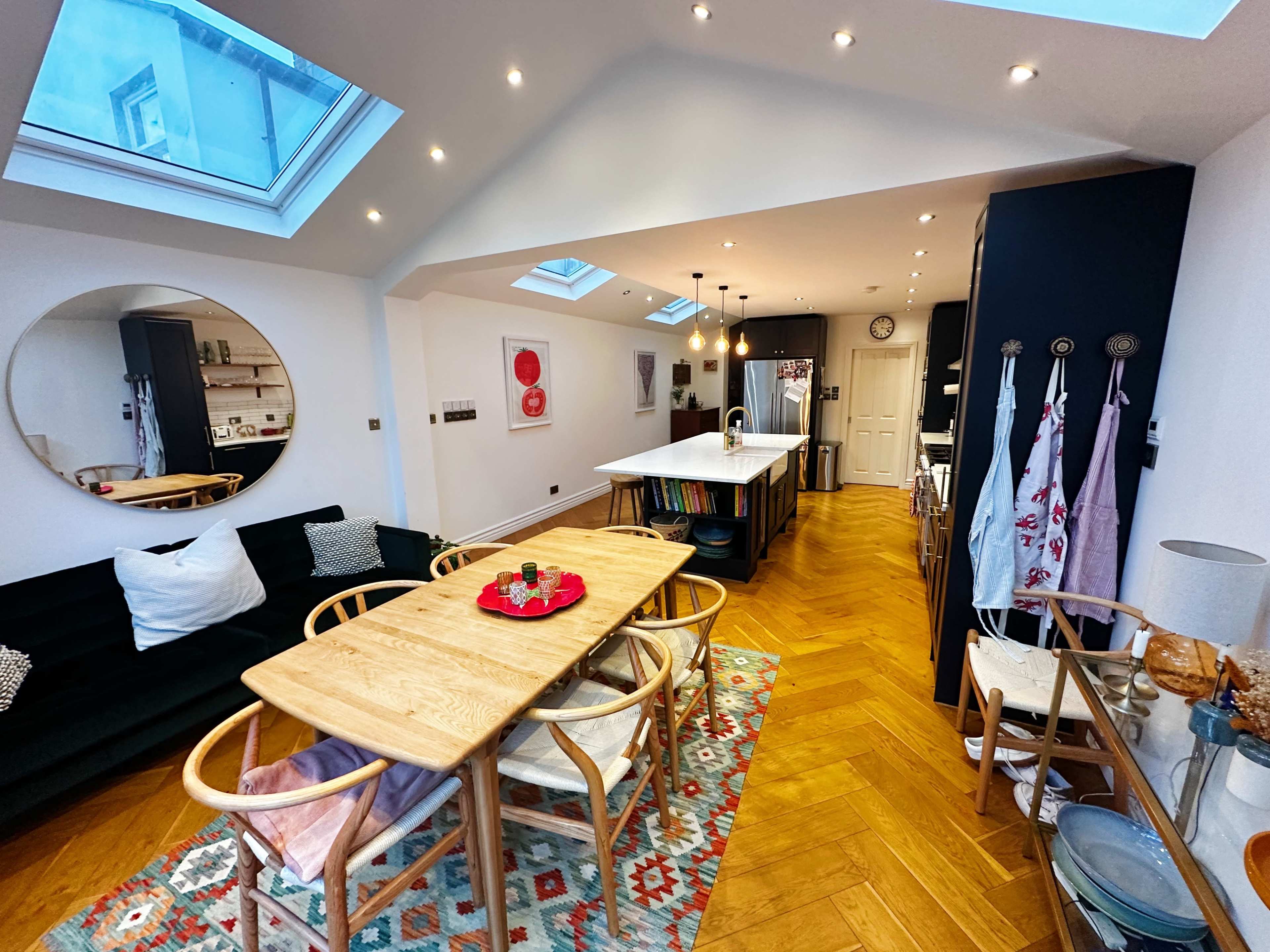 The image shows a modern kitchen and dining area with a wooden table, chairs, and a large skylight.