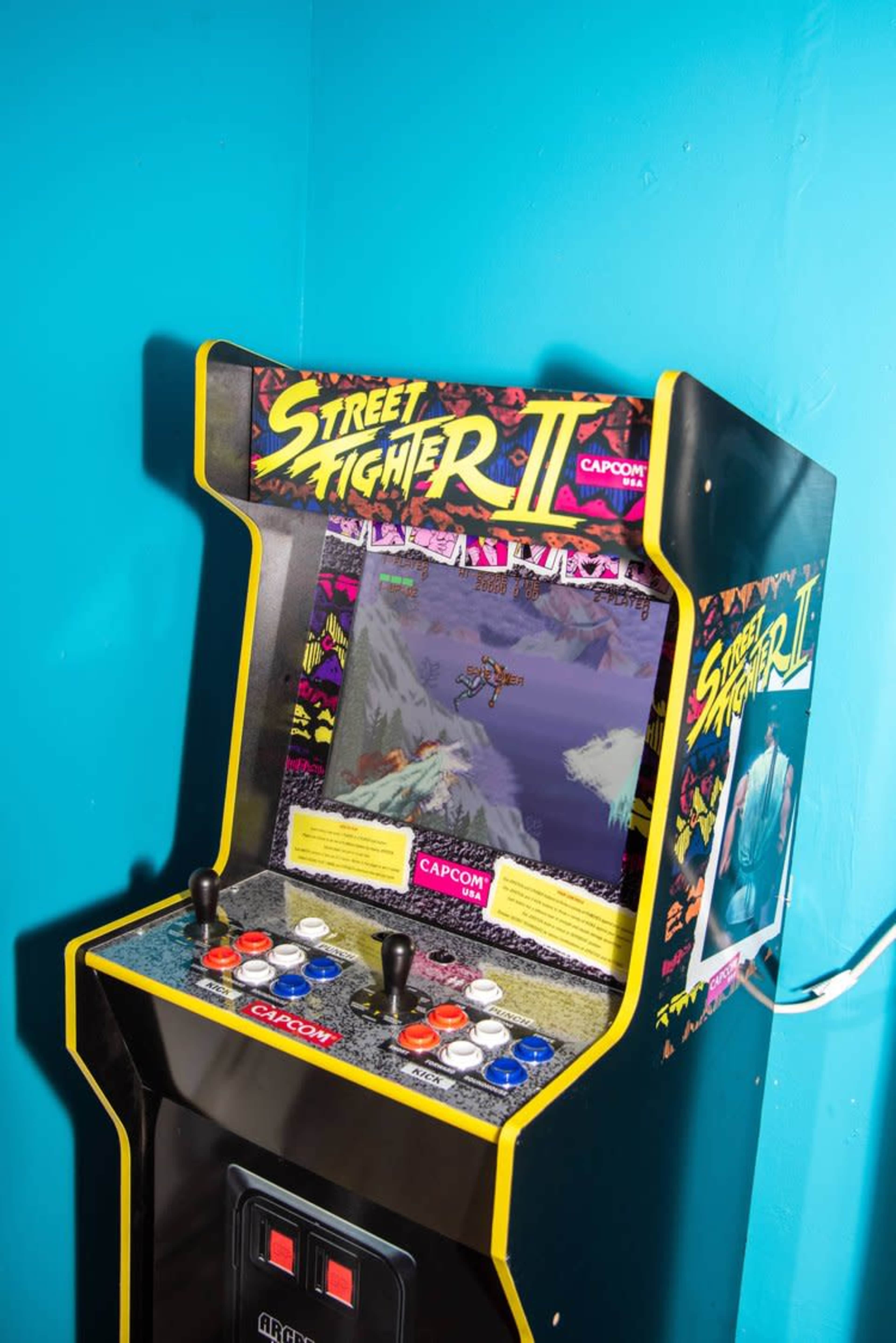 An arcade machine for the game "Street Fighter II" stands against a blue wall.