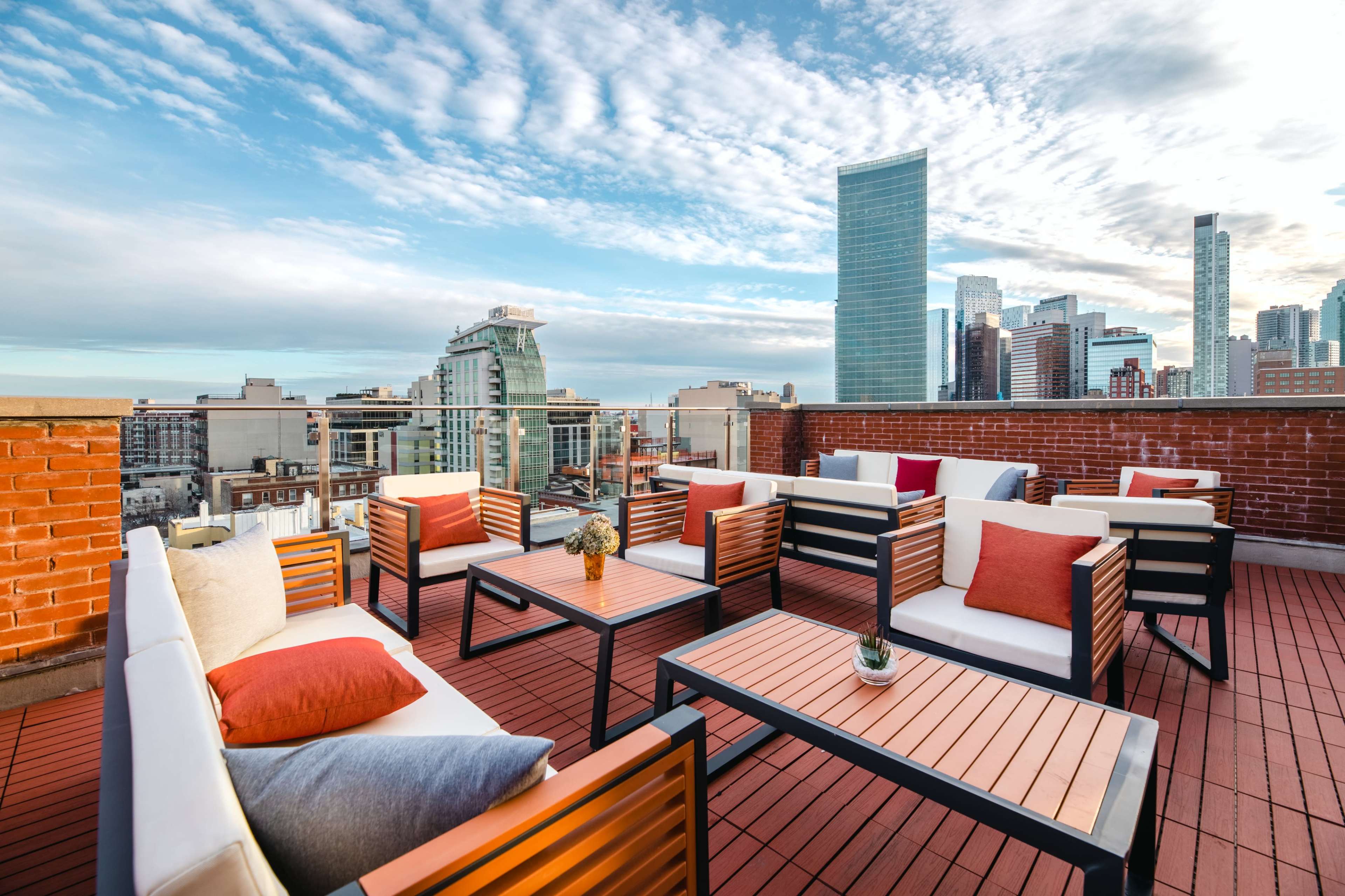 A rooftop terrace featuring modern seating arrangements with tables, overlooking a city skyline under a partly cloudy sky.