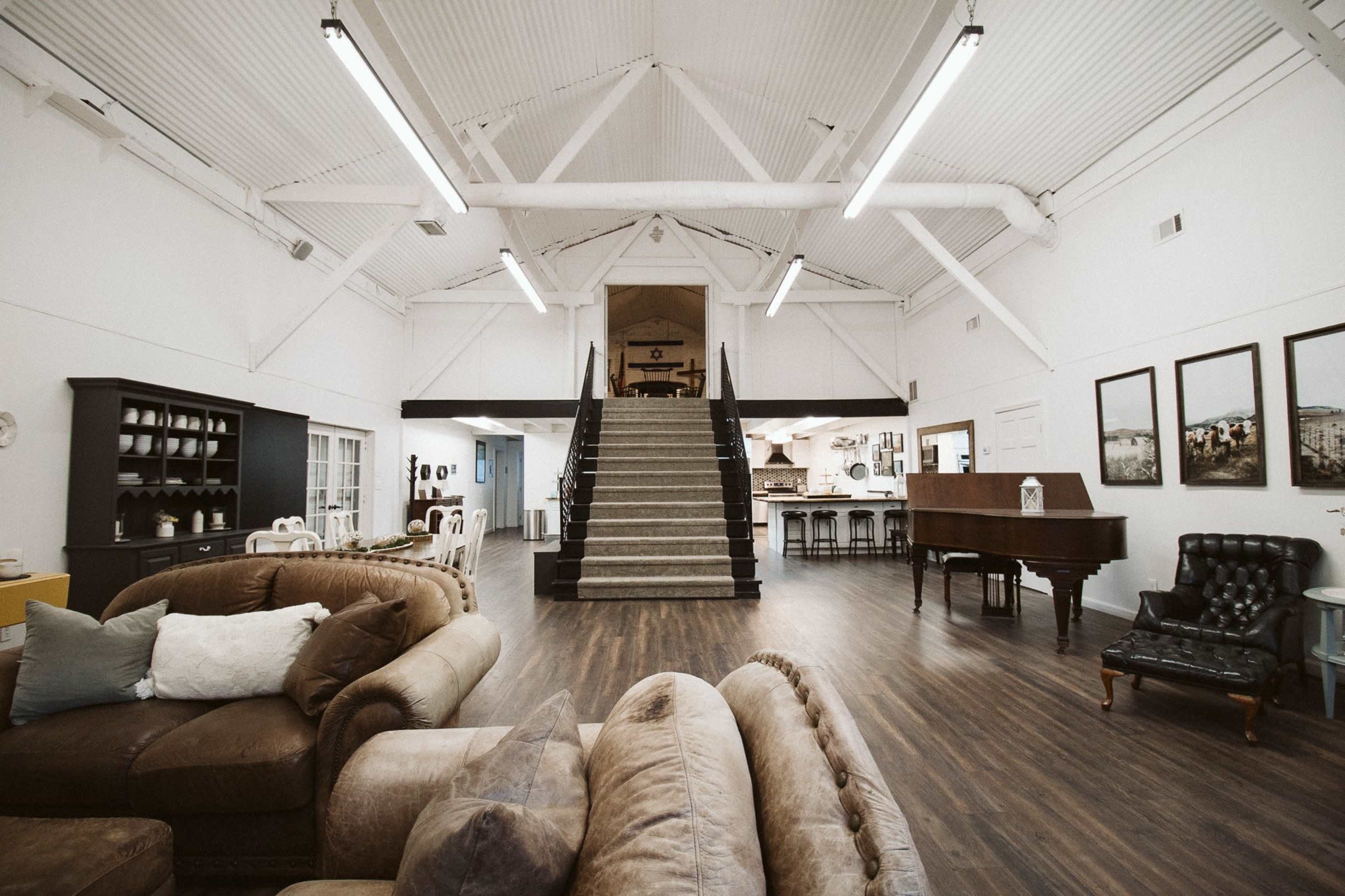 A spacious, open interior with high ceilings, featuring a staircase in the center that leads to an upper level, surrounded by various furniture arrangements.
