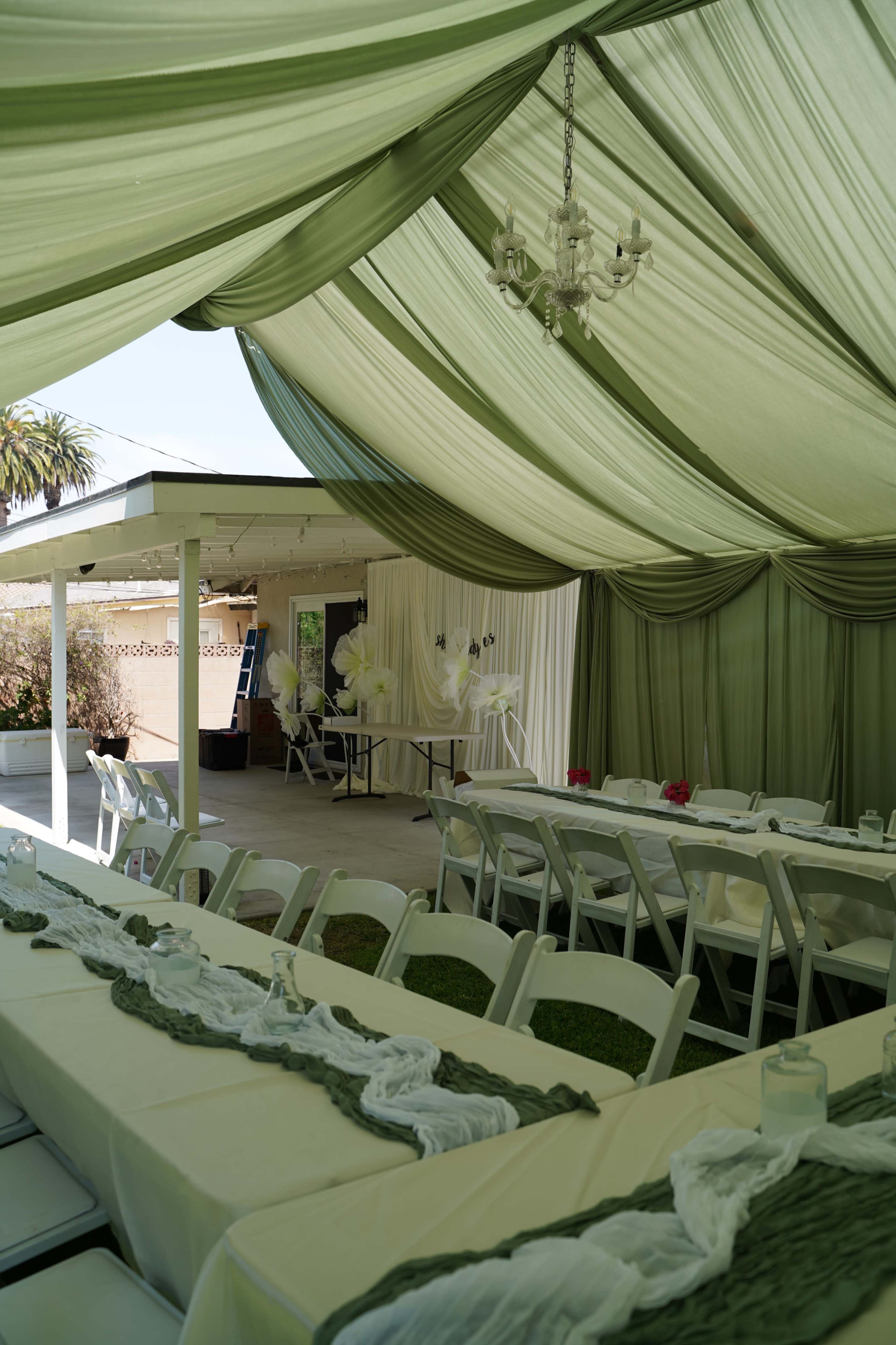 The image shows a decorated outdoor event space with long tables set up under a canopy of draped green fabric.