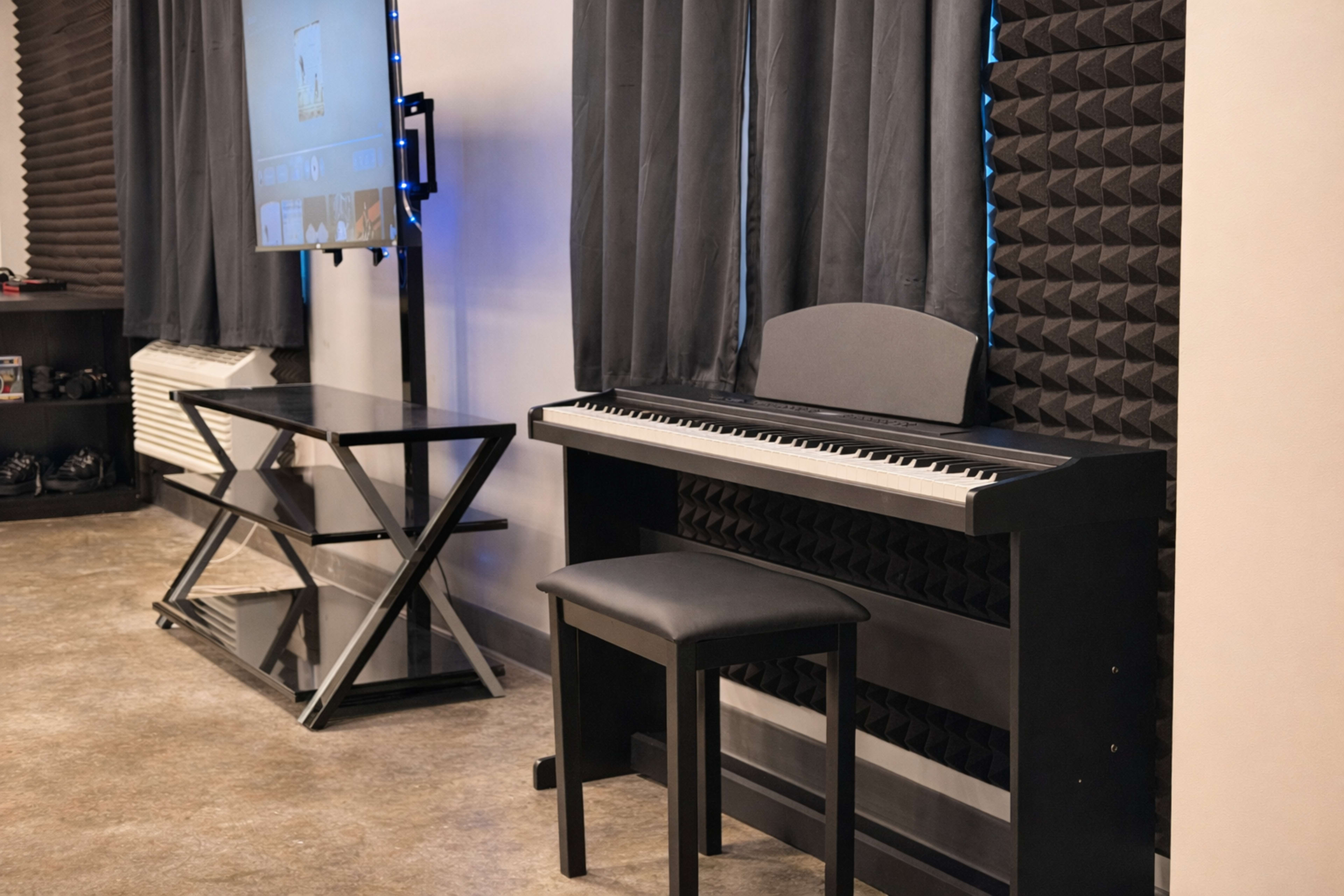 The image depicts a black digital piano with a stool, positioned beside a modern glass table and a large screen mounted on the wall in a room with dark curtains and soundproofing panels.