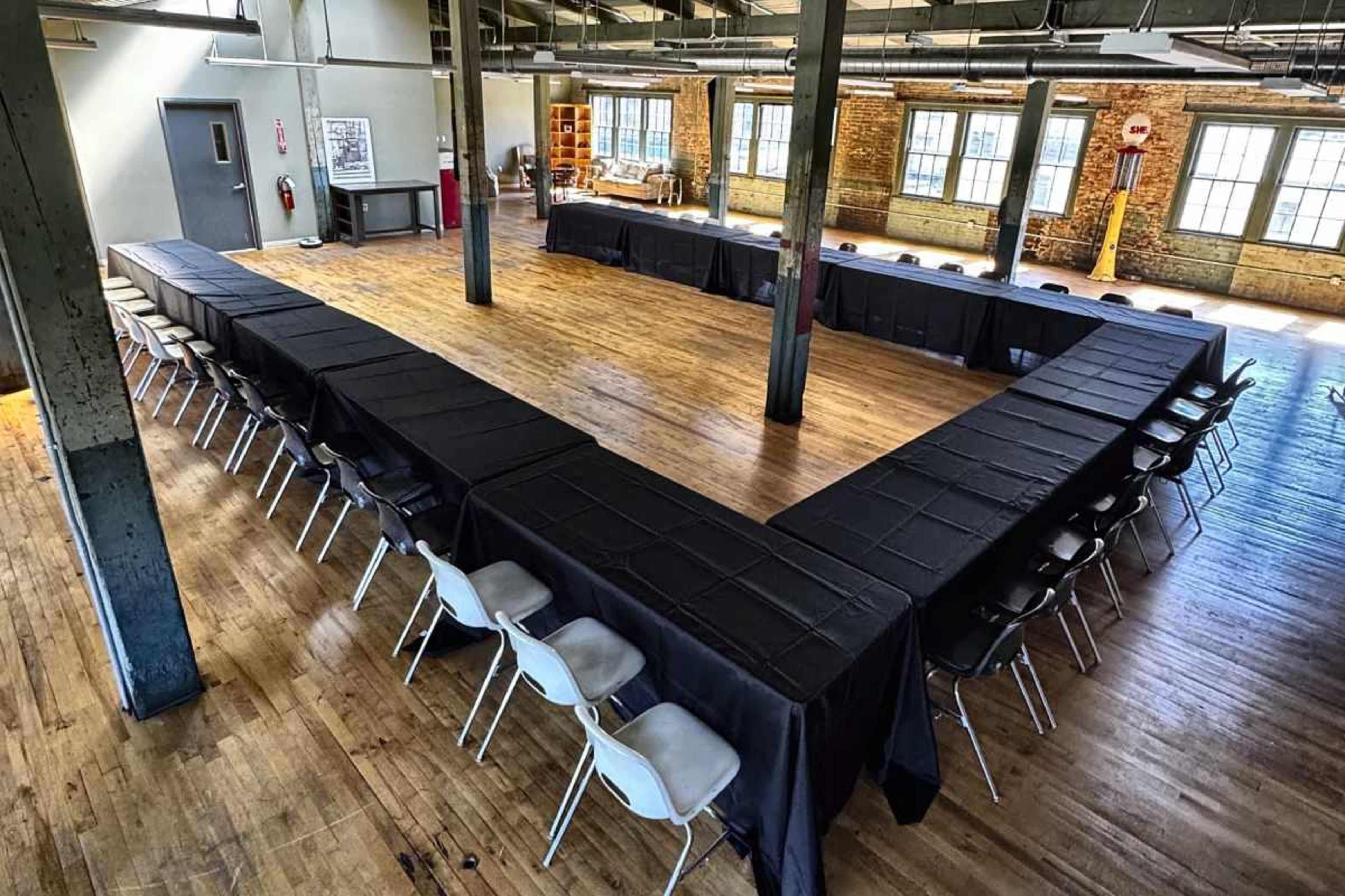 A large, rectangular room features a series of black-covered tables arranged in a U-shape, surrounded by white chairs on a hardwood floor.