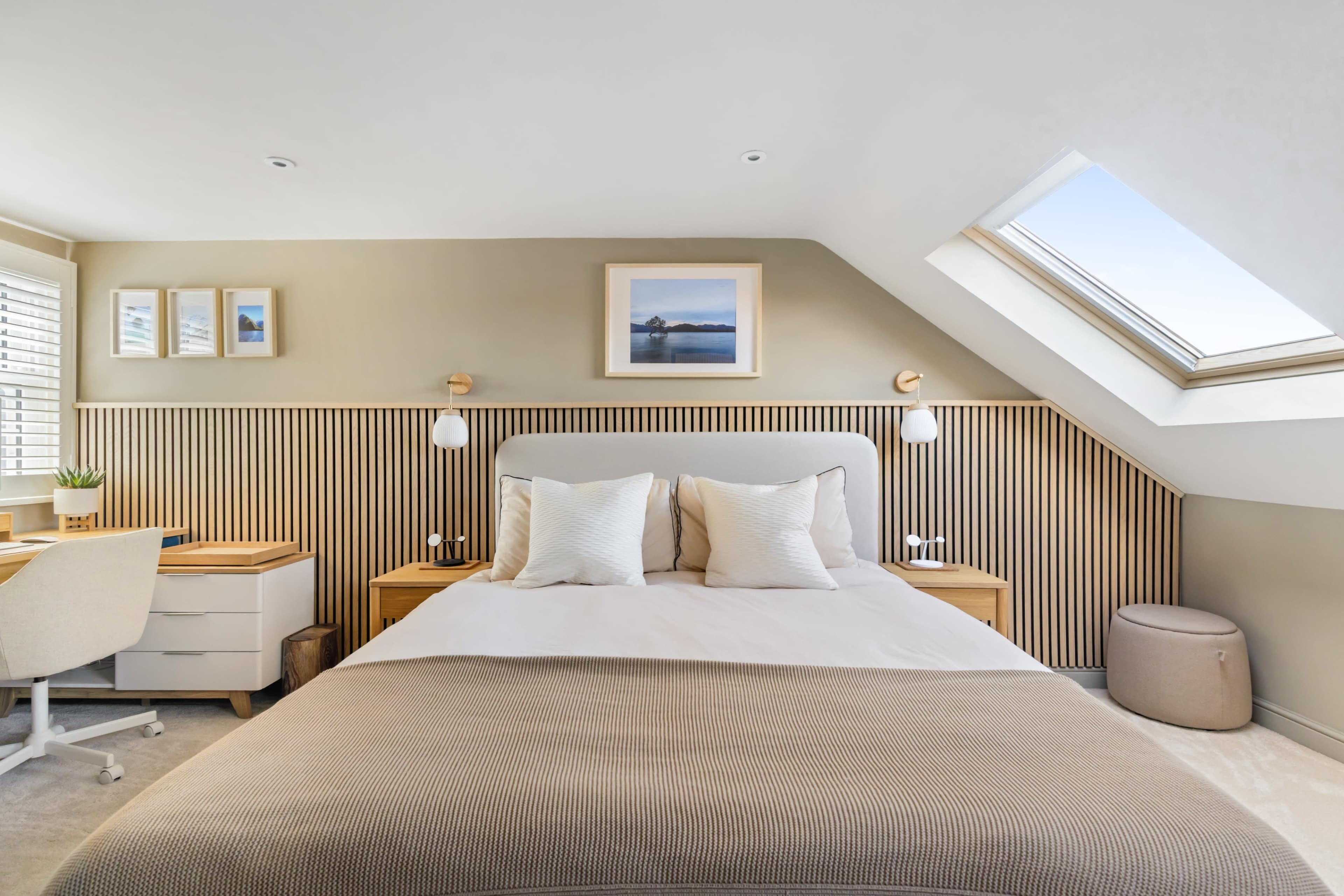 The image depicts a modern bedroom with a bed, two nightstands, and a desk under a sloped ceiling, featuring natural light from a skylight.