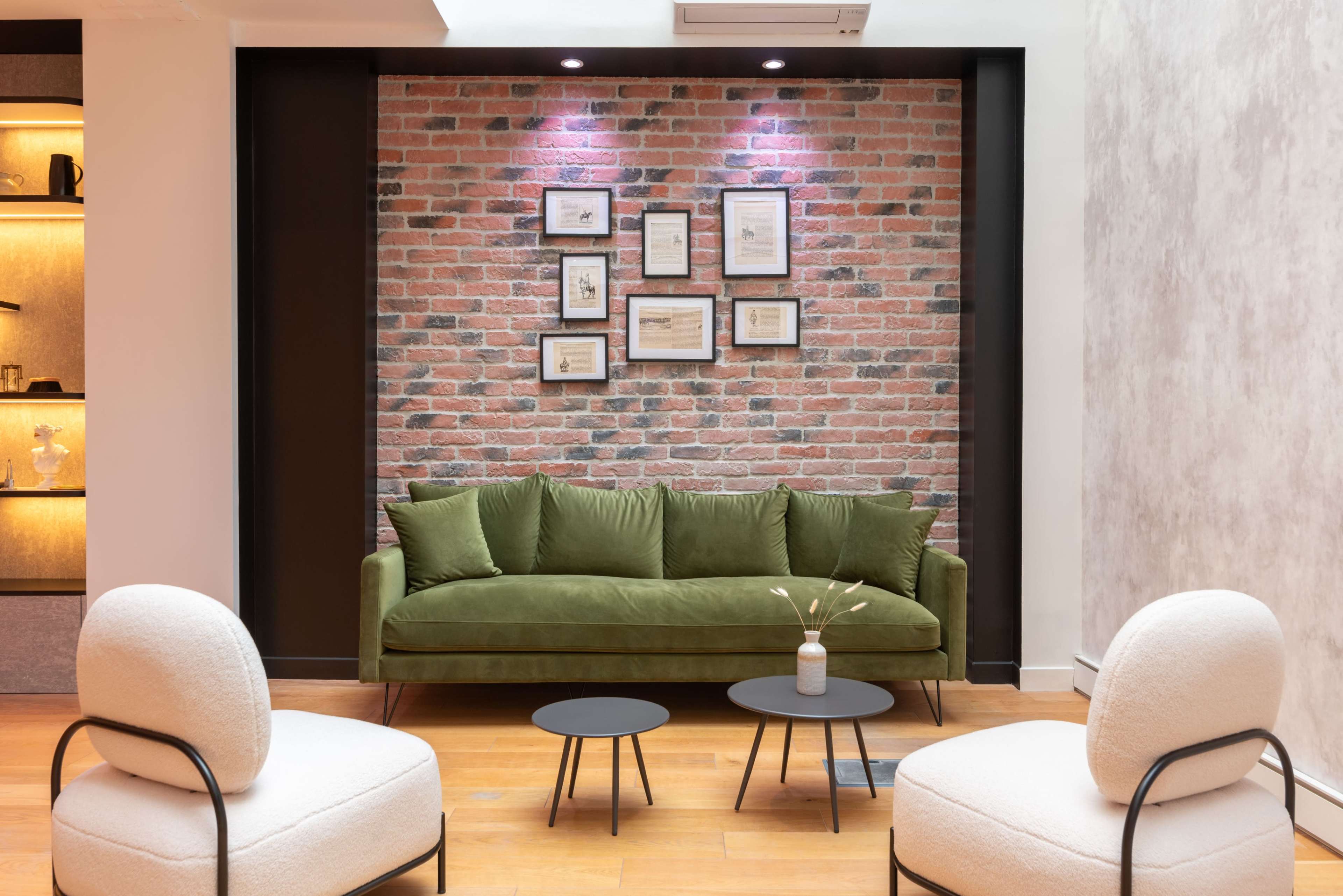 A green sofa is positioned against a brick wall adorned with framed artworks, with two round tables and two upholstered chairs in front.