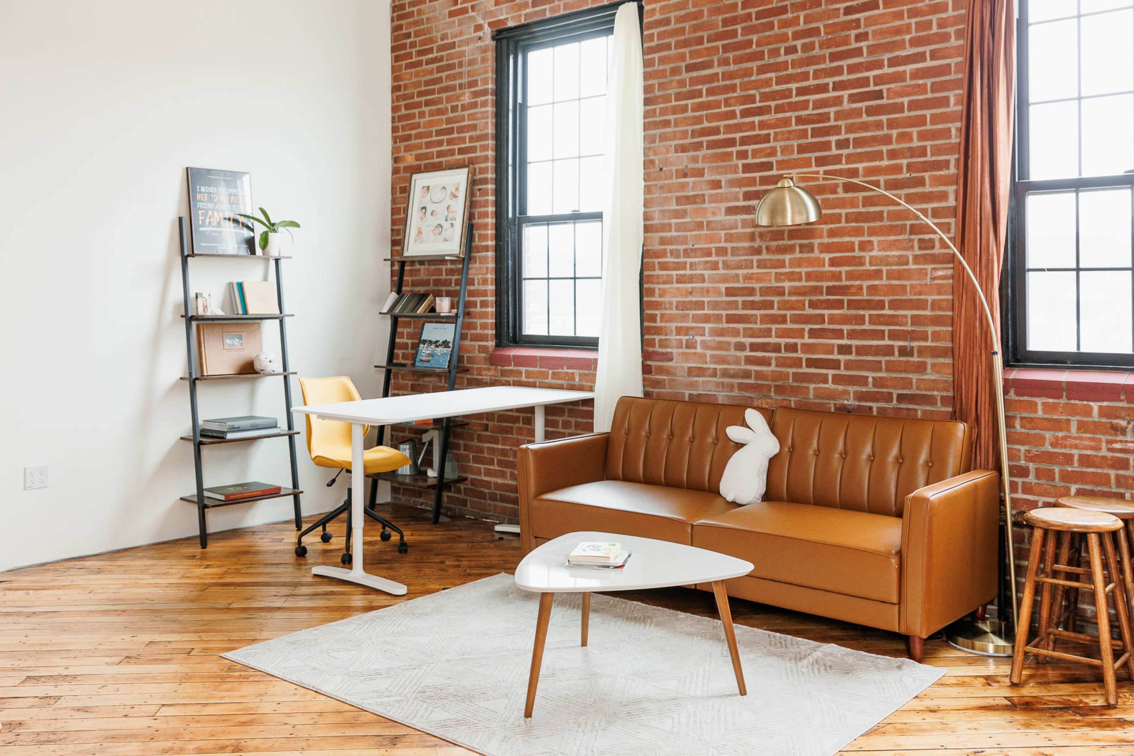 A modern living space featuring a brown leather sofa, a white coffee table, a yellow office chair, and a bookshelf adorned with various items against a brick wall.