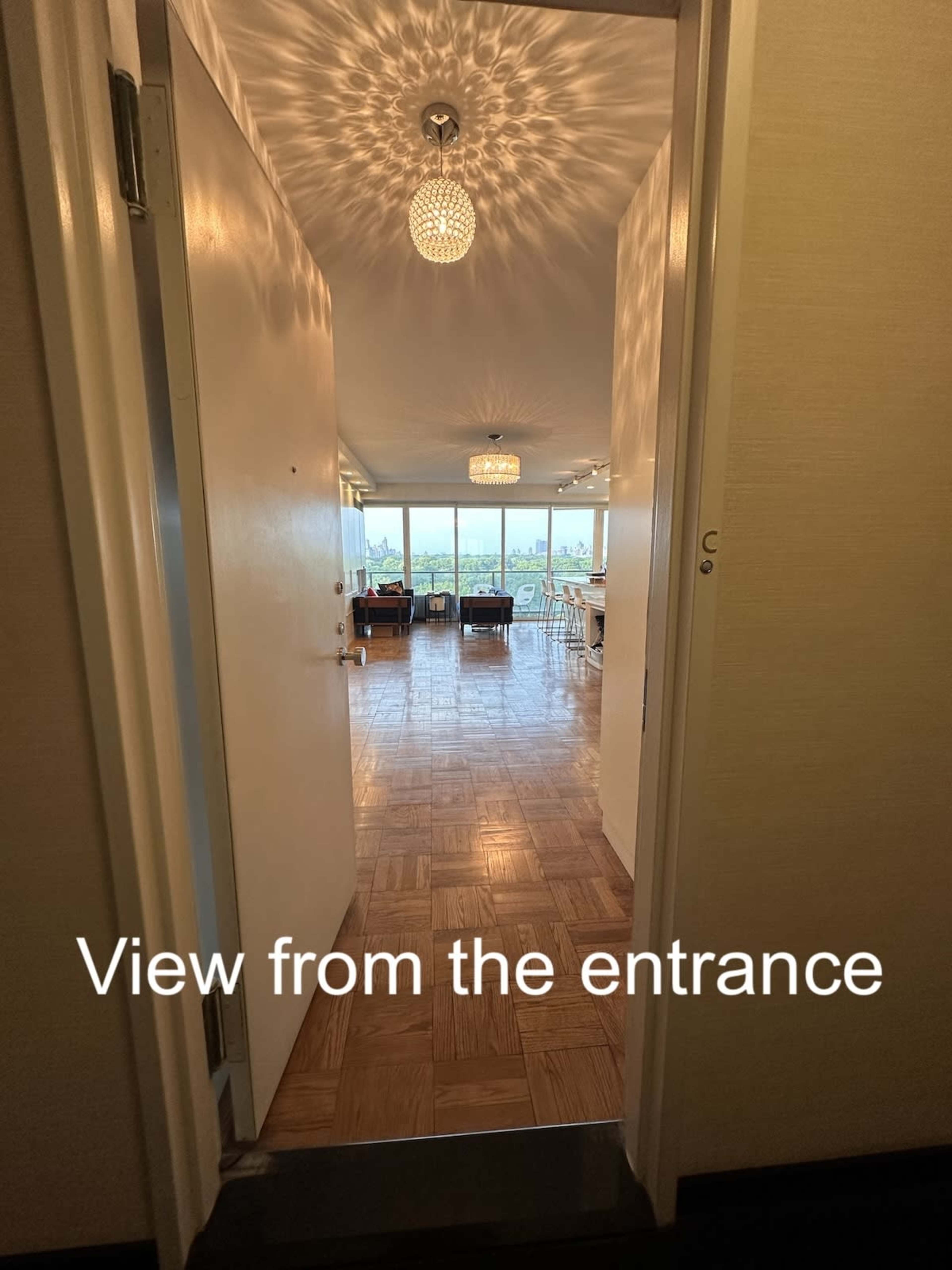 The image shows a view through an open door into a spacious room featuring wooden flooring and large windows with a cityscape visible outside.