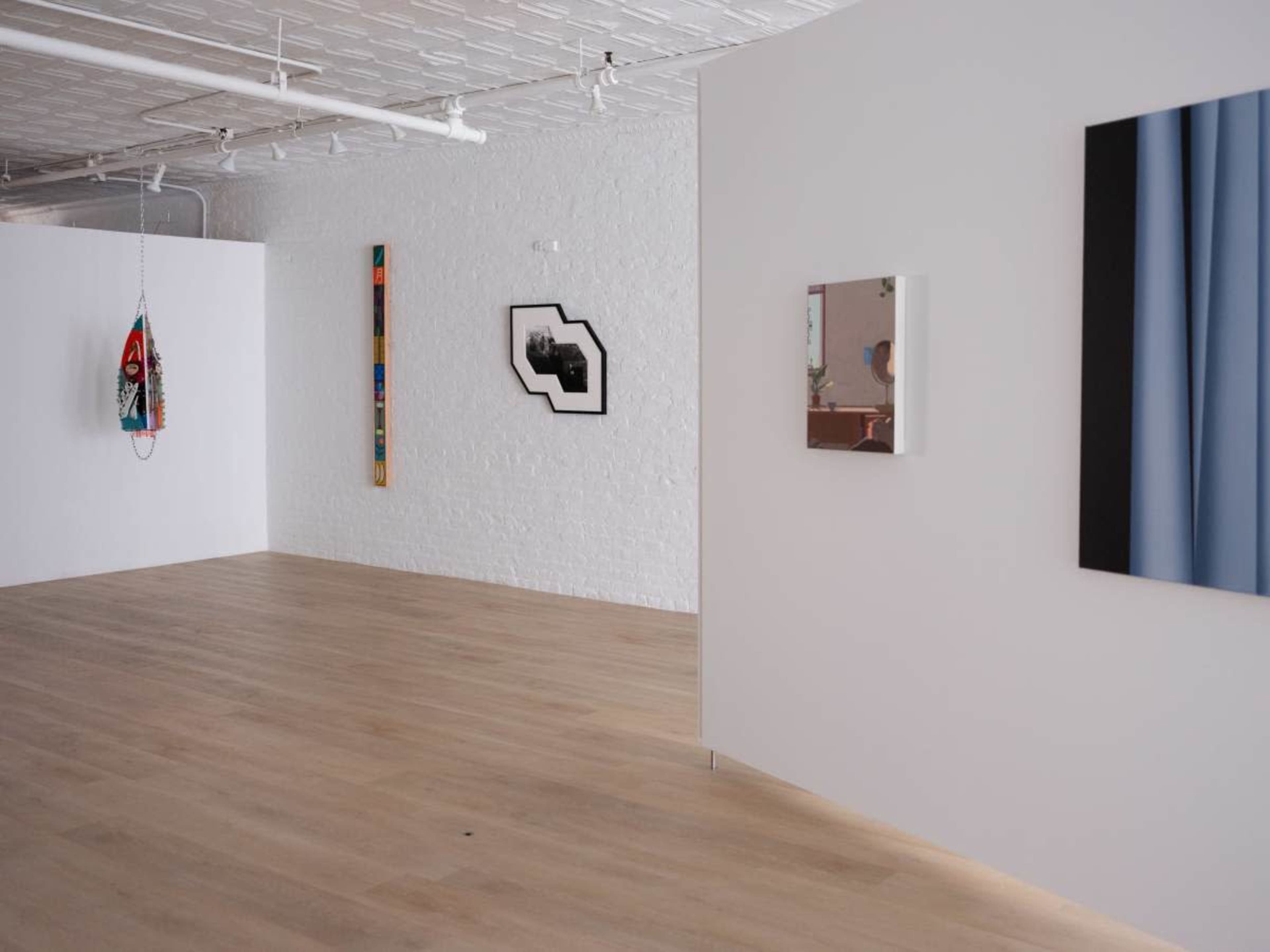 The image shows a contemporary art gallery with white walls displaying various artworks, including framed pieces and a hanging sculpture, set in a spacious room with wooden flooring.