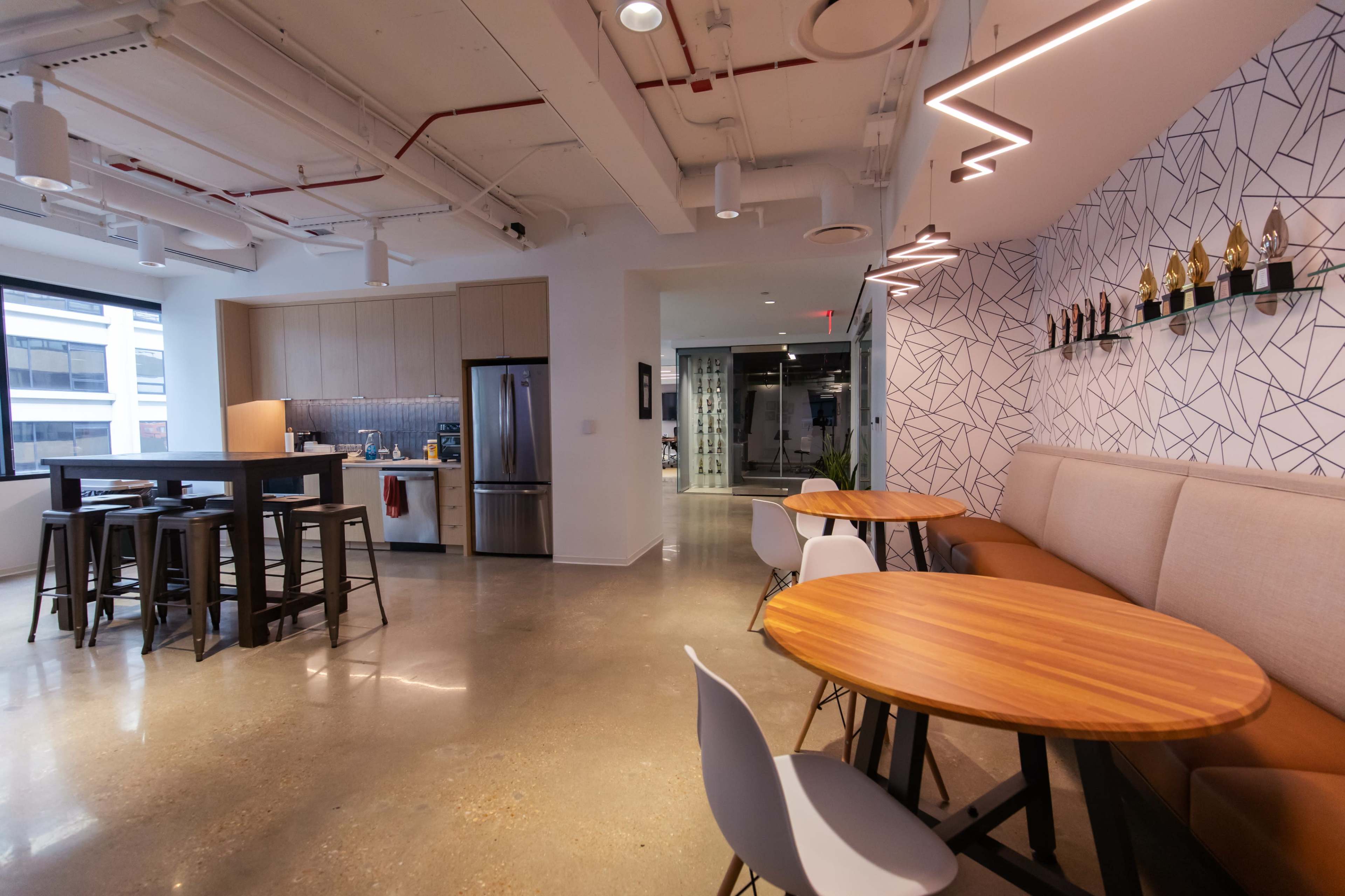 A modern office space featuring a kitchenette, dining area with wooden tables, and a wall displaying several trophies.
