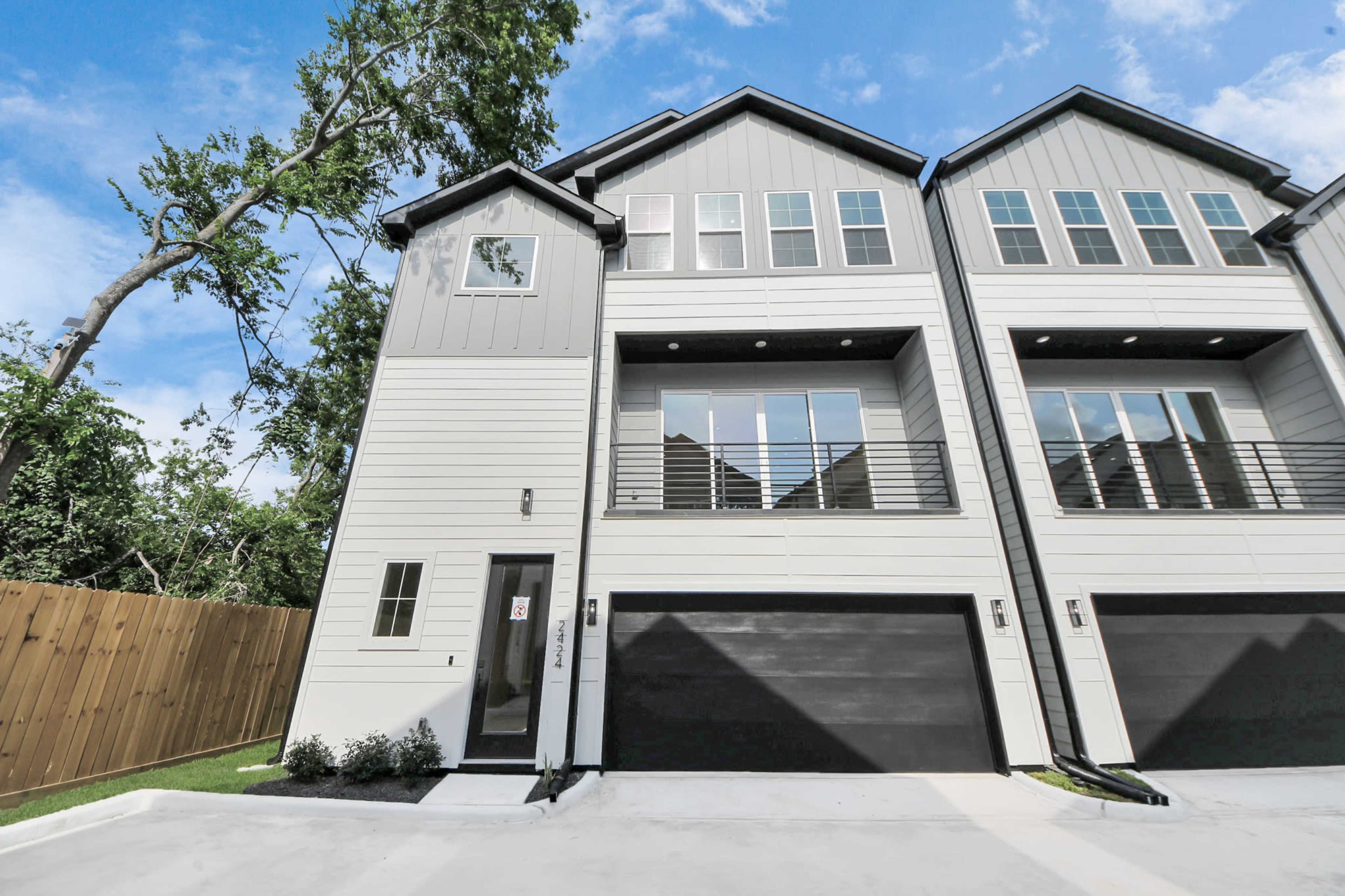 The image shows a modern two-story duplex with a light-colored exterior and large windows, situated next to a wooden fence and trees.