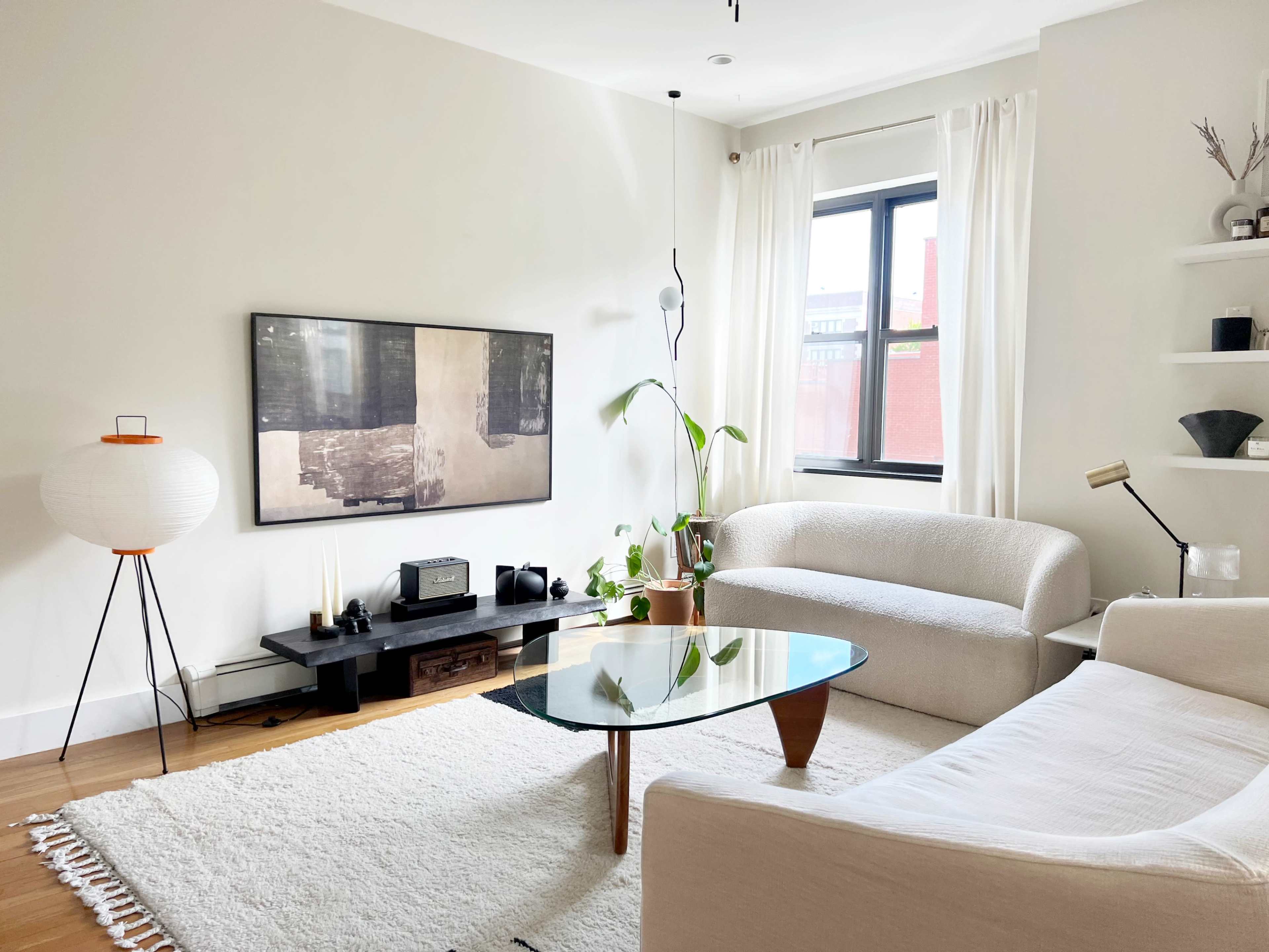 A modern living room with a white sofa, a glass coffee table, a flat-screen TV mounted on the wall, and a potted plant near a window.