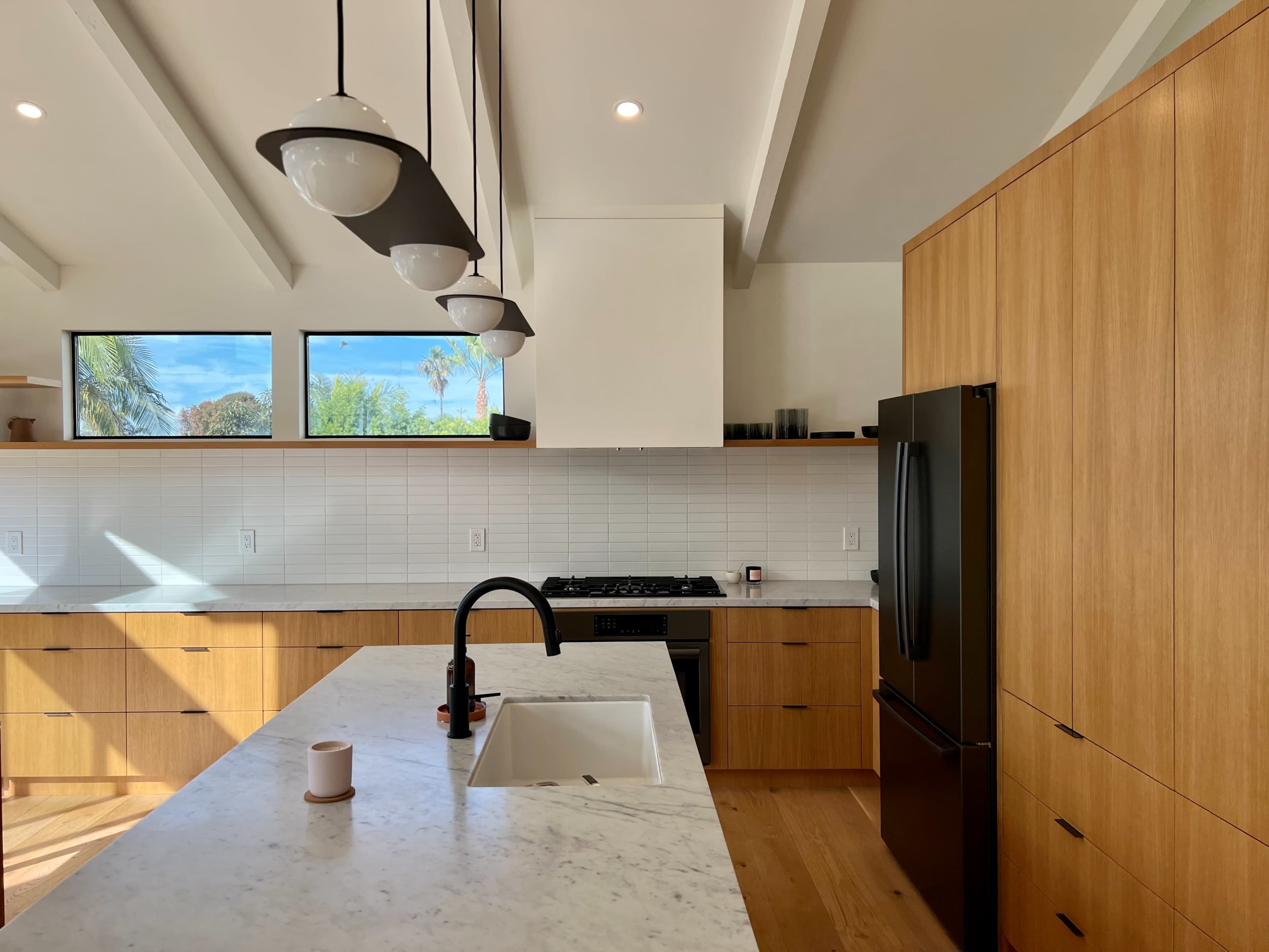 A modern kitchen with wooden cabinetry, a marble countertop, a black refrigerator, and large windows allowing natural light to enter.