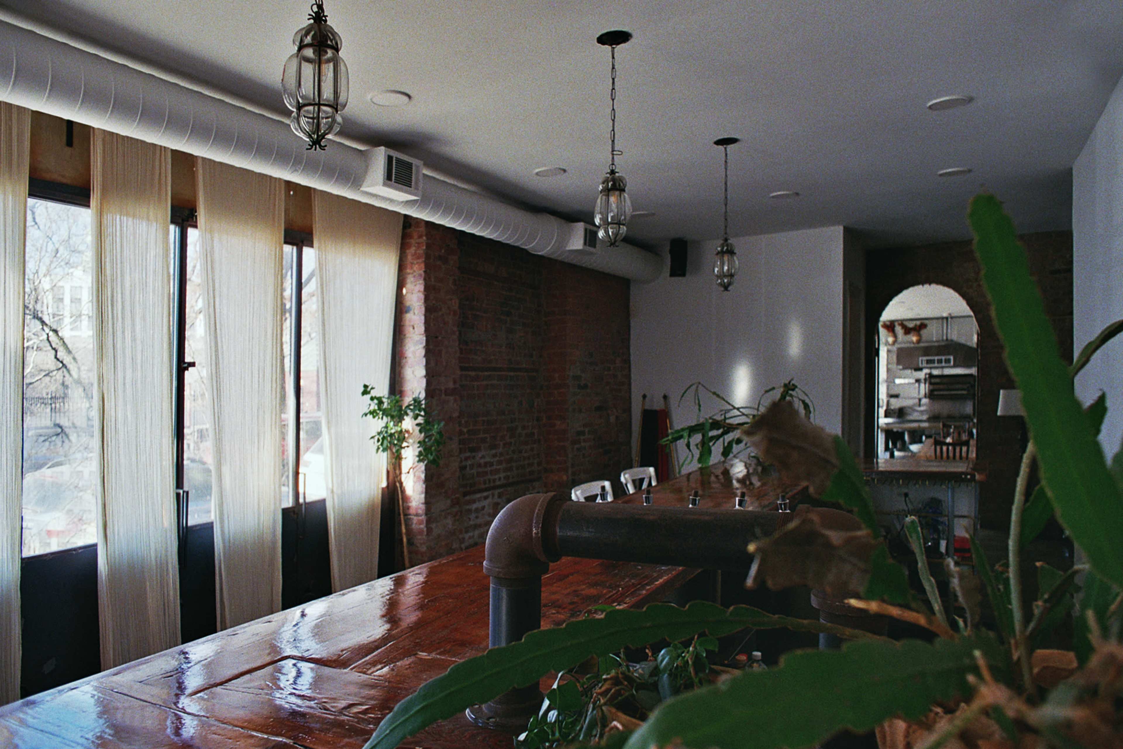 The image shows a spacious interior of a cafe or restaurant featuring a long wooden table, large windows with sheer curtains, and hanging light fixtures.