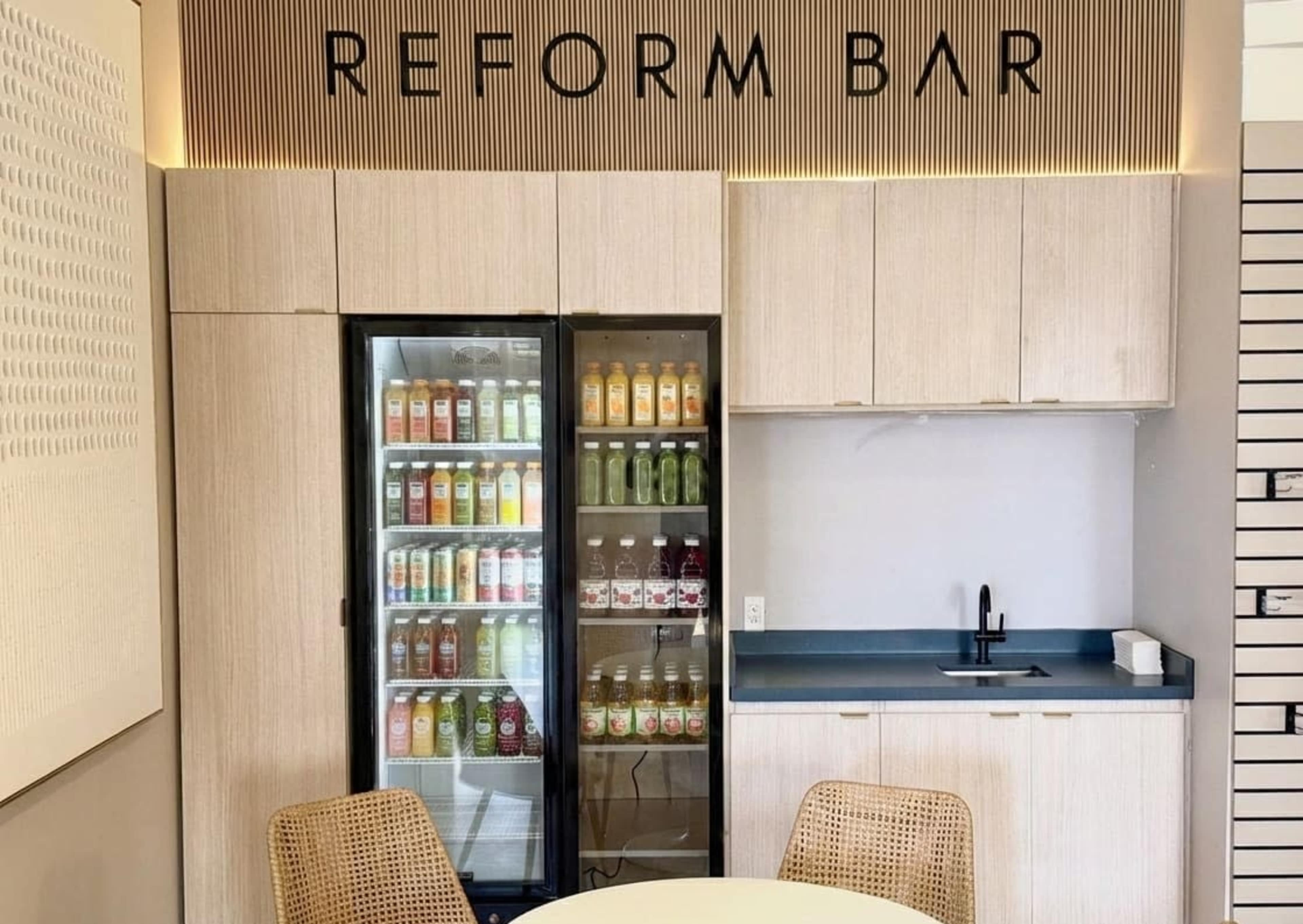 The image shows a modern interior featuring a beverage refrigerator filled with various drinks, a countertop, and the words "REFORM BAR" displayed above.