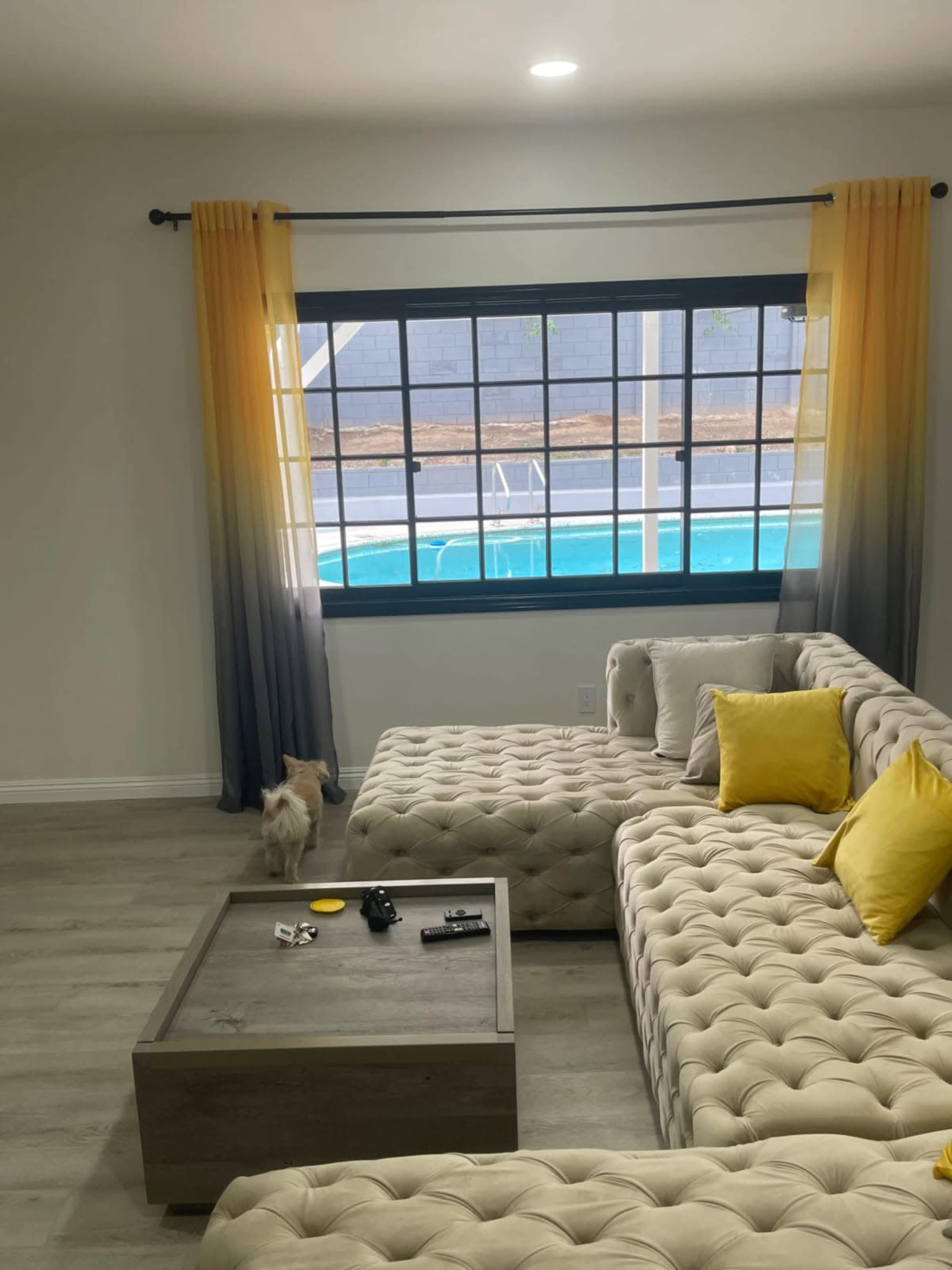 A light-filled living room features a tufted sofa with yellow cushions, a wooden coffee table, and a large window overlooking a swimming pool.