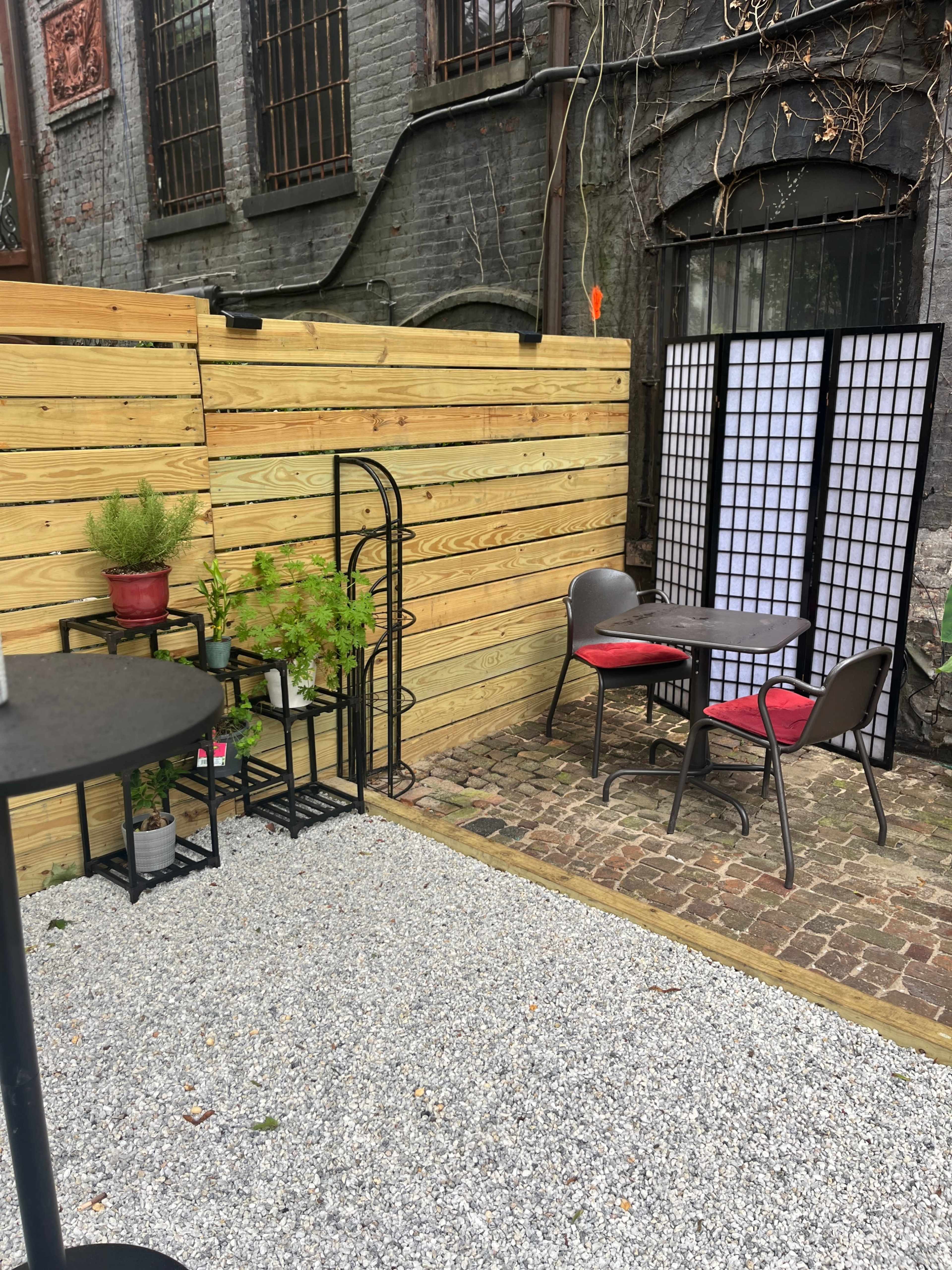 A small outdoor seating area features a gravel surface with two chairs and a table, surrounded by wooden fences and plants in pots.