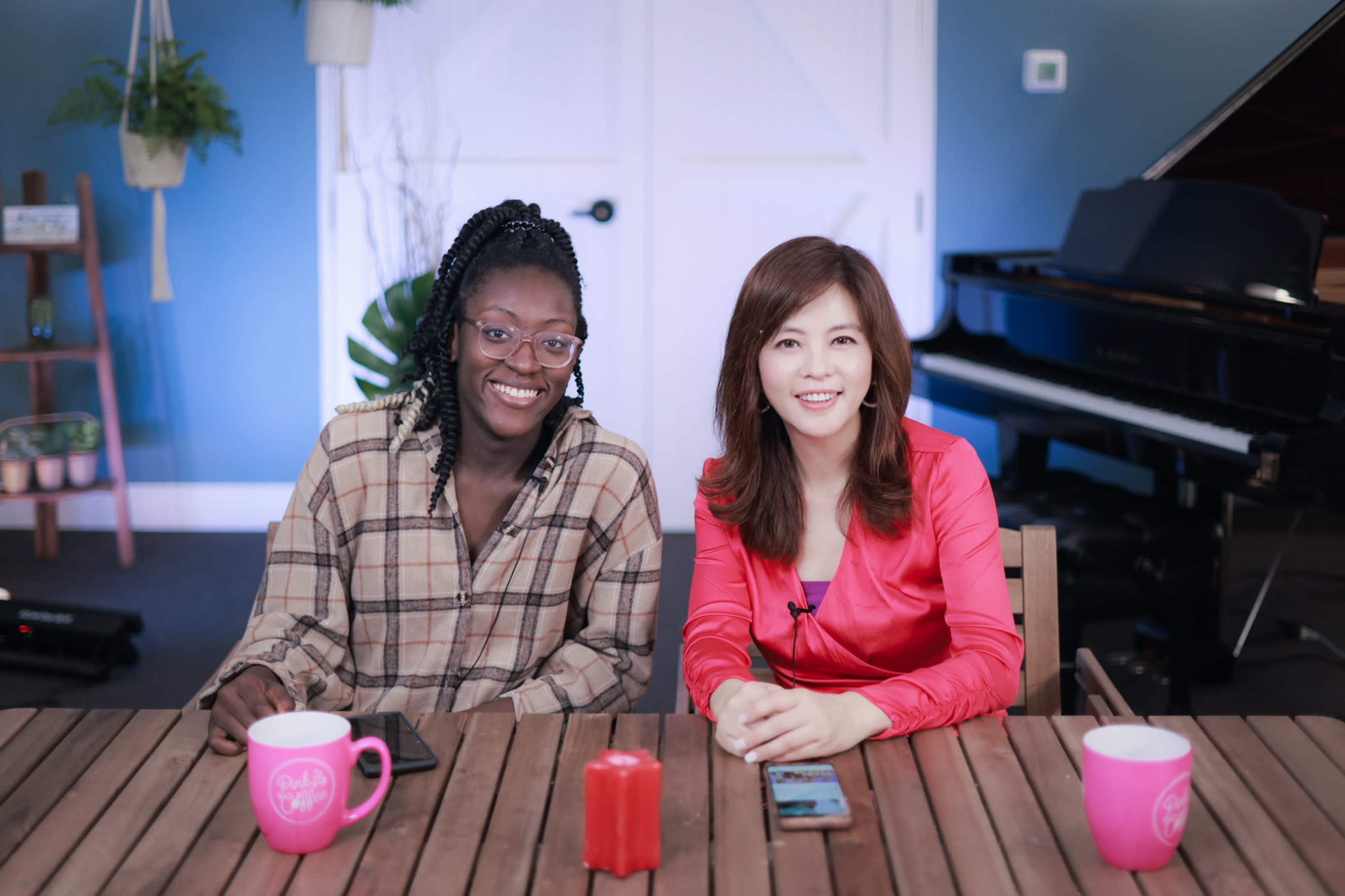 Two women sit at a wooden table, smiling, with coffee mugs in front of them, and a piano in the background.