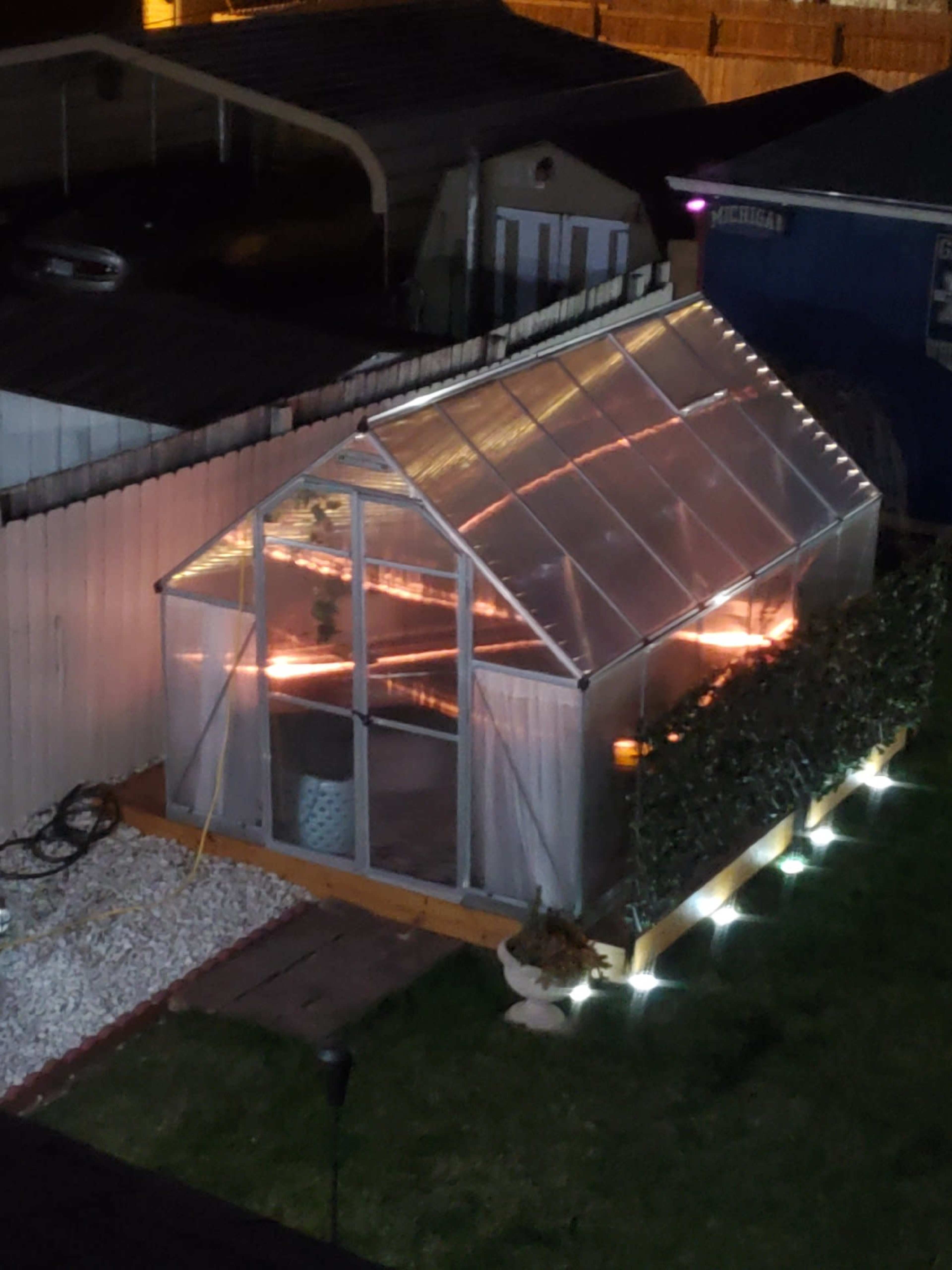 A greenhouse with clear panels and a wooden base is illuminated at night, surrounded by a neatly trimmed hedge and gravel landscaping.