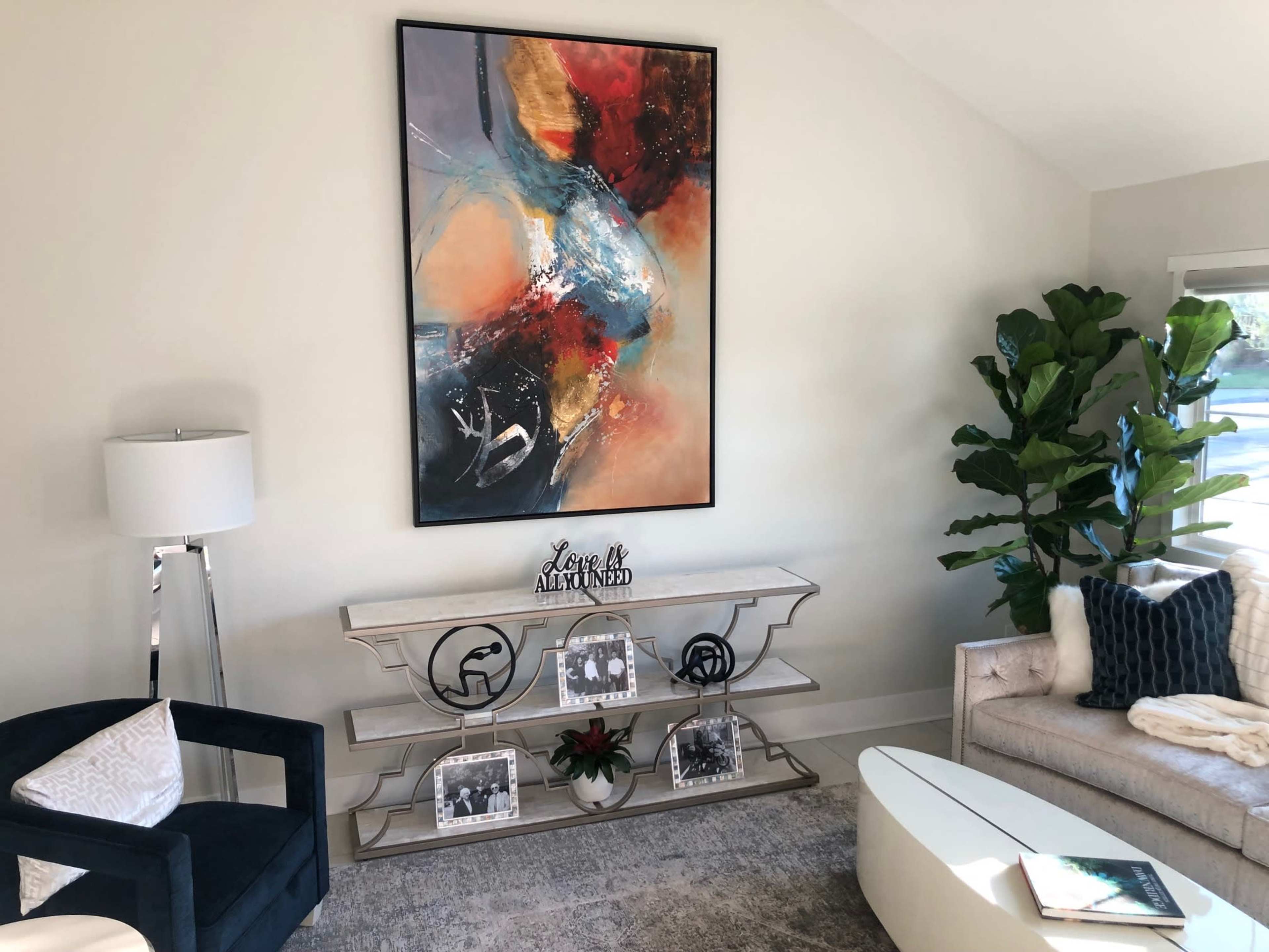The image features a modern living room with a large abstract painting hanging above a console table, accompanied by a comfortable sofa, armchair, and a decorative plant.
