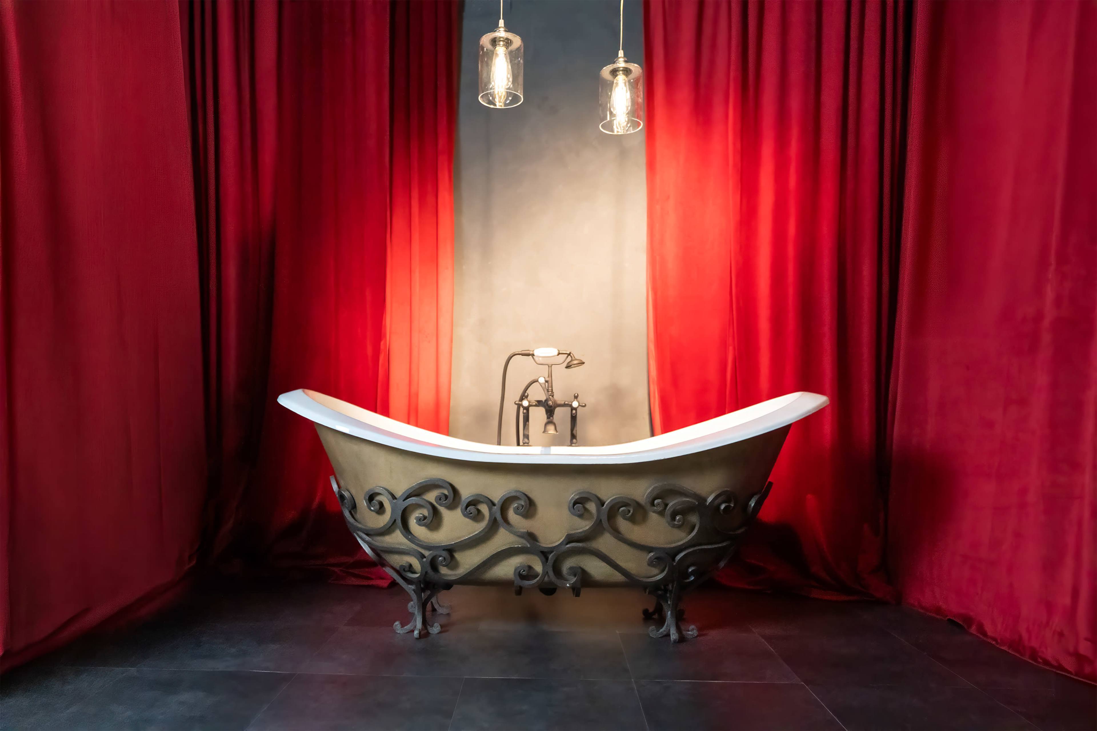A vintage-style bathtub with ornate metal detailing is positioned against a backdrop of dark red curtains and illuminated by pendant lights.