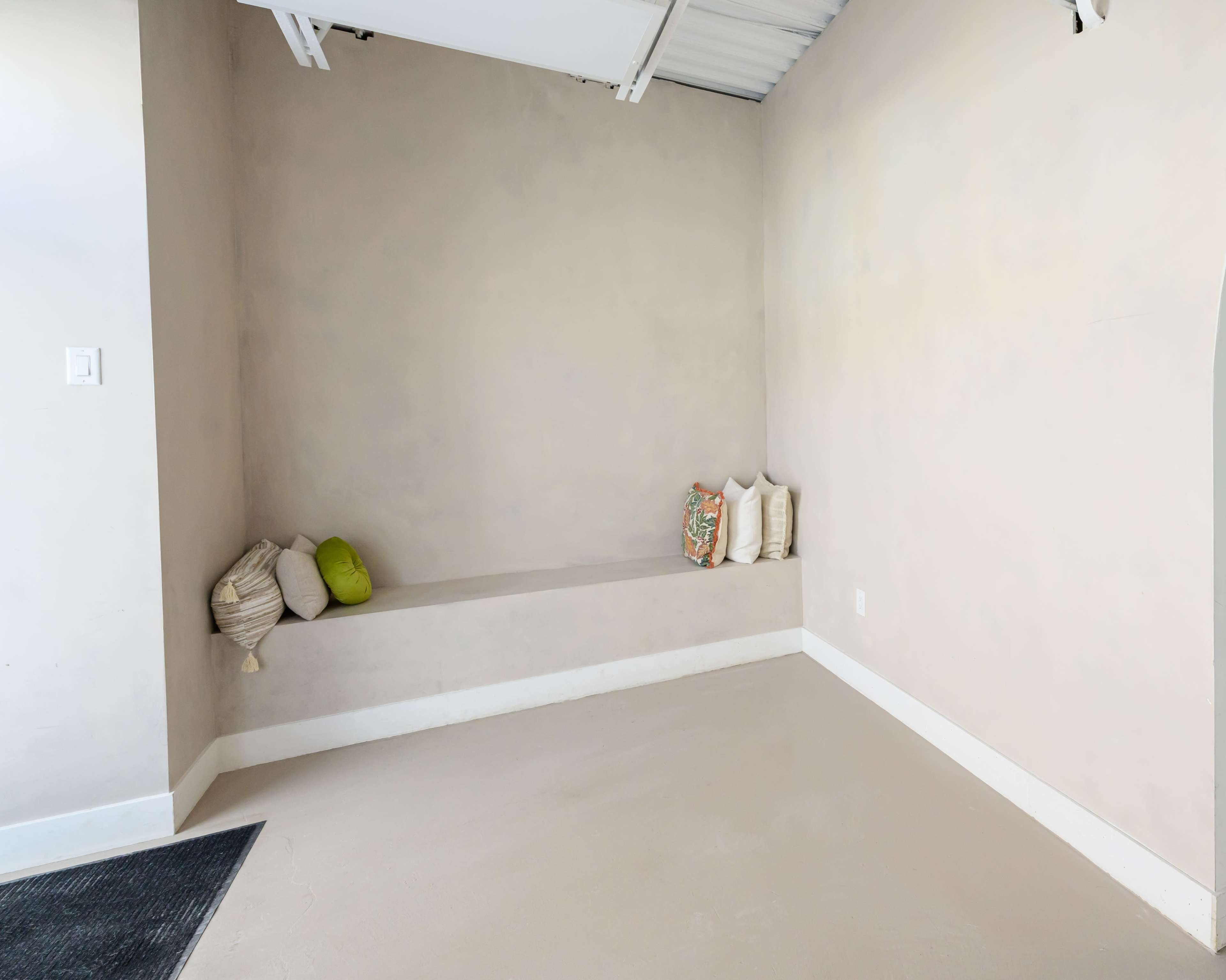 The image shows a minimalist room with a smooth, light-colored wall, a built-in bench with a few decorative pillows, and a bare floor.