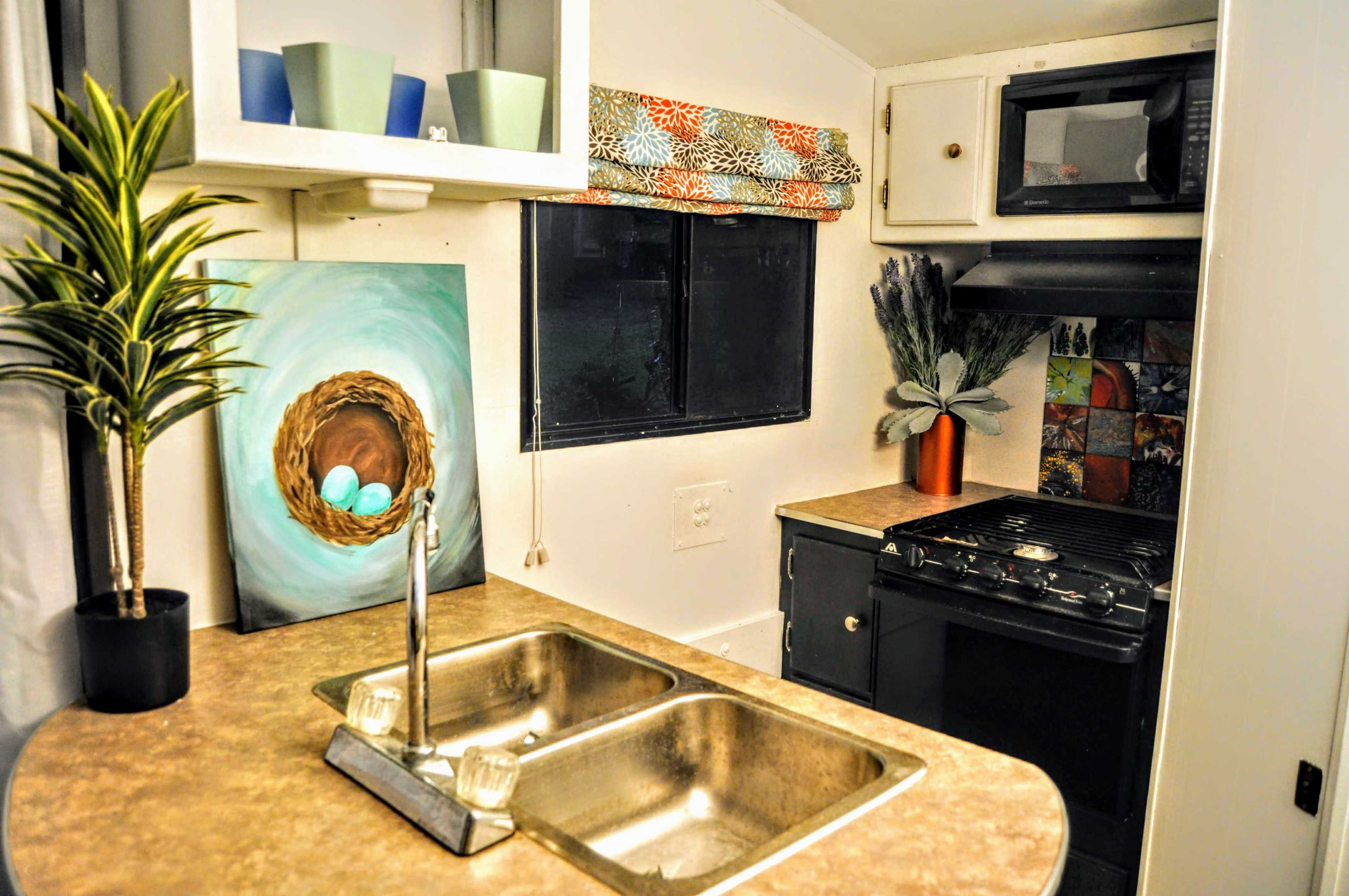 The image shows a compact kitchen area with a double sink, stove, and a window adorned with colorful fabric, alongside decorative elements like a plant and an artwork of a bird's nest with eggs.