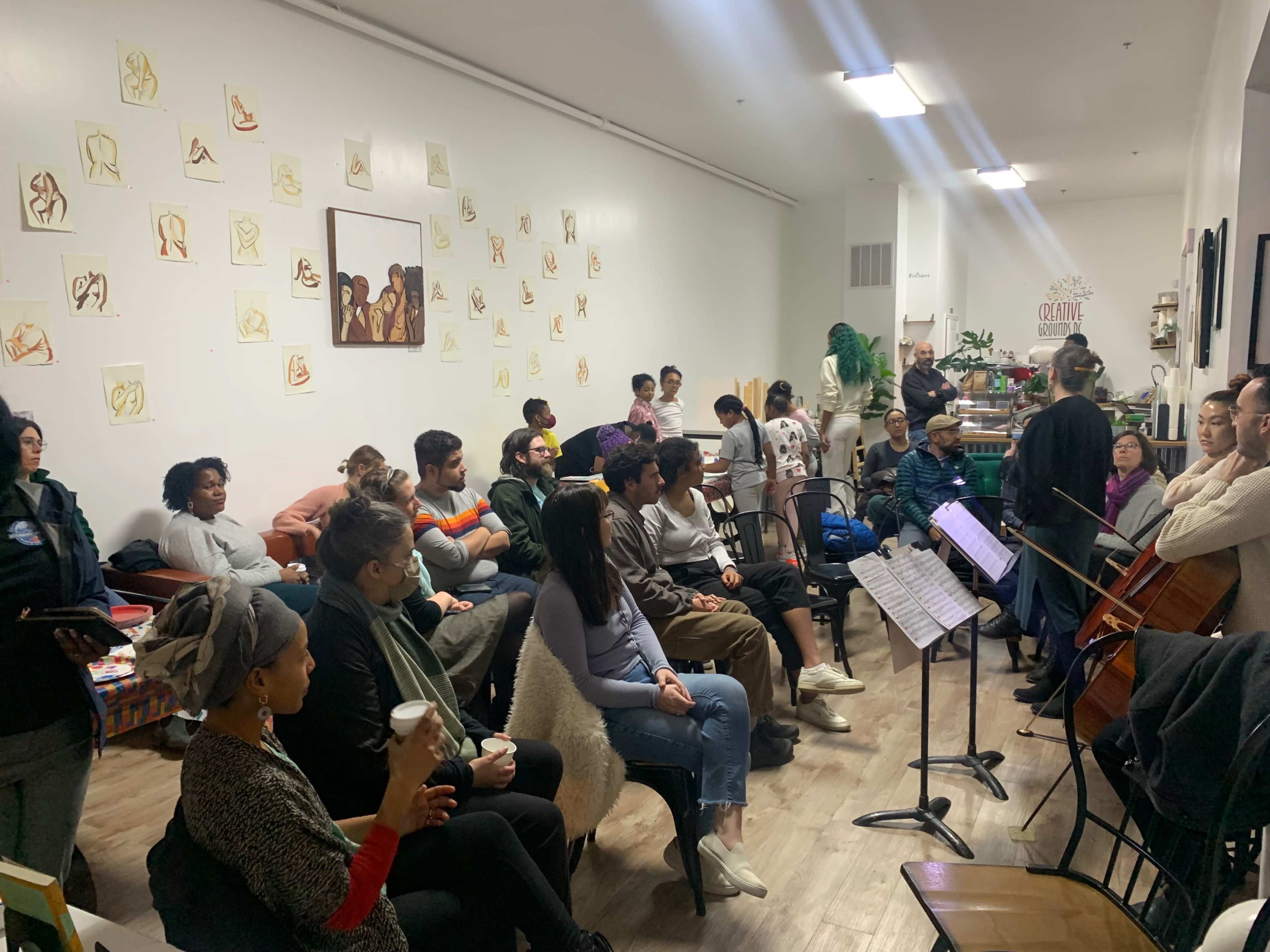 A diverse group of people is seated in a small, well-lit room, listening attentively to musicians performing with a cello and other instruments while art sketches adorn the walls.