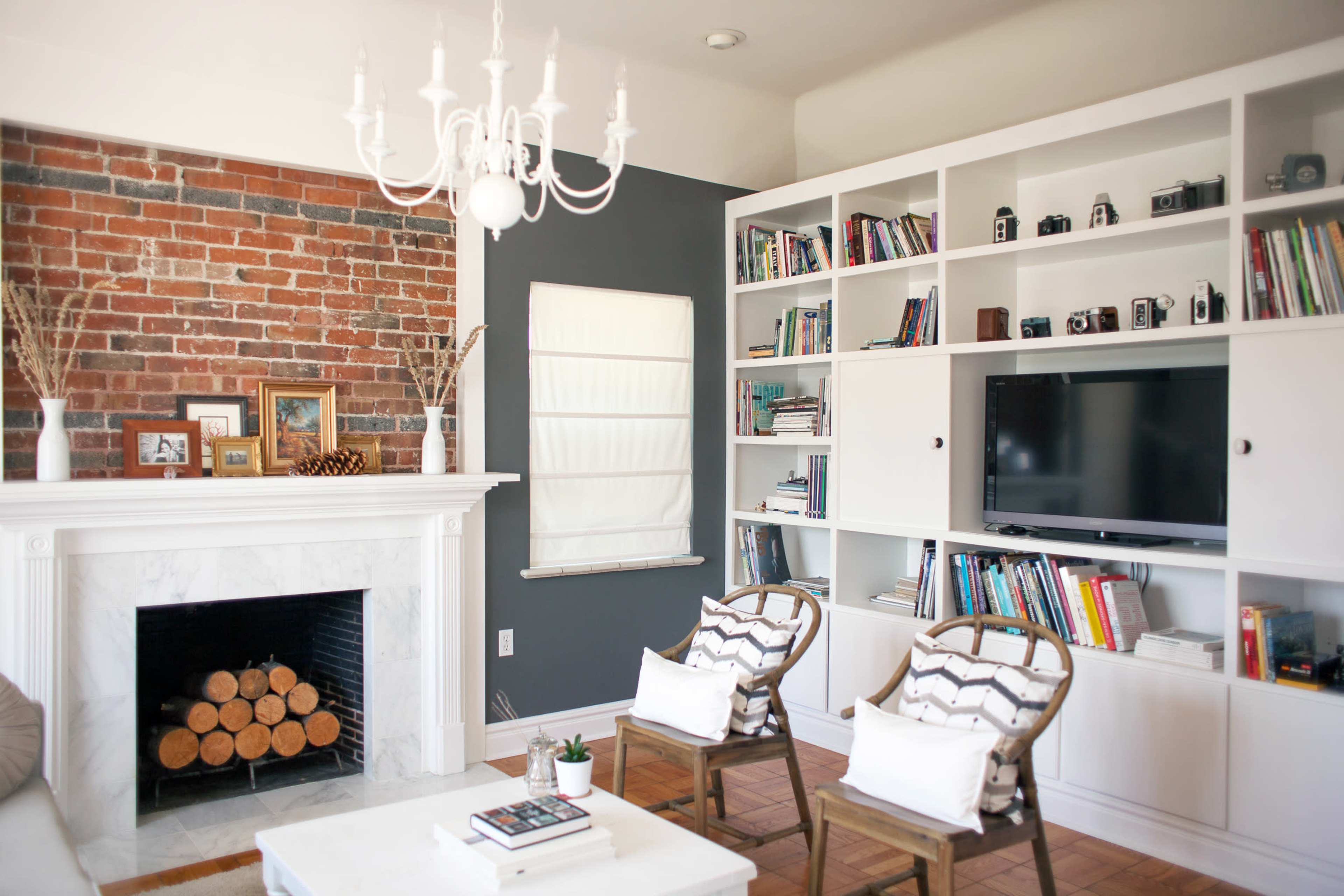 A living room featuring a brick accent wall, a white fireplace with stacked logs, two wooden chairs, a large bookshelf filled with books and cameras, and a television positioned on a white cabinet.