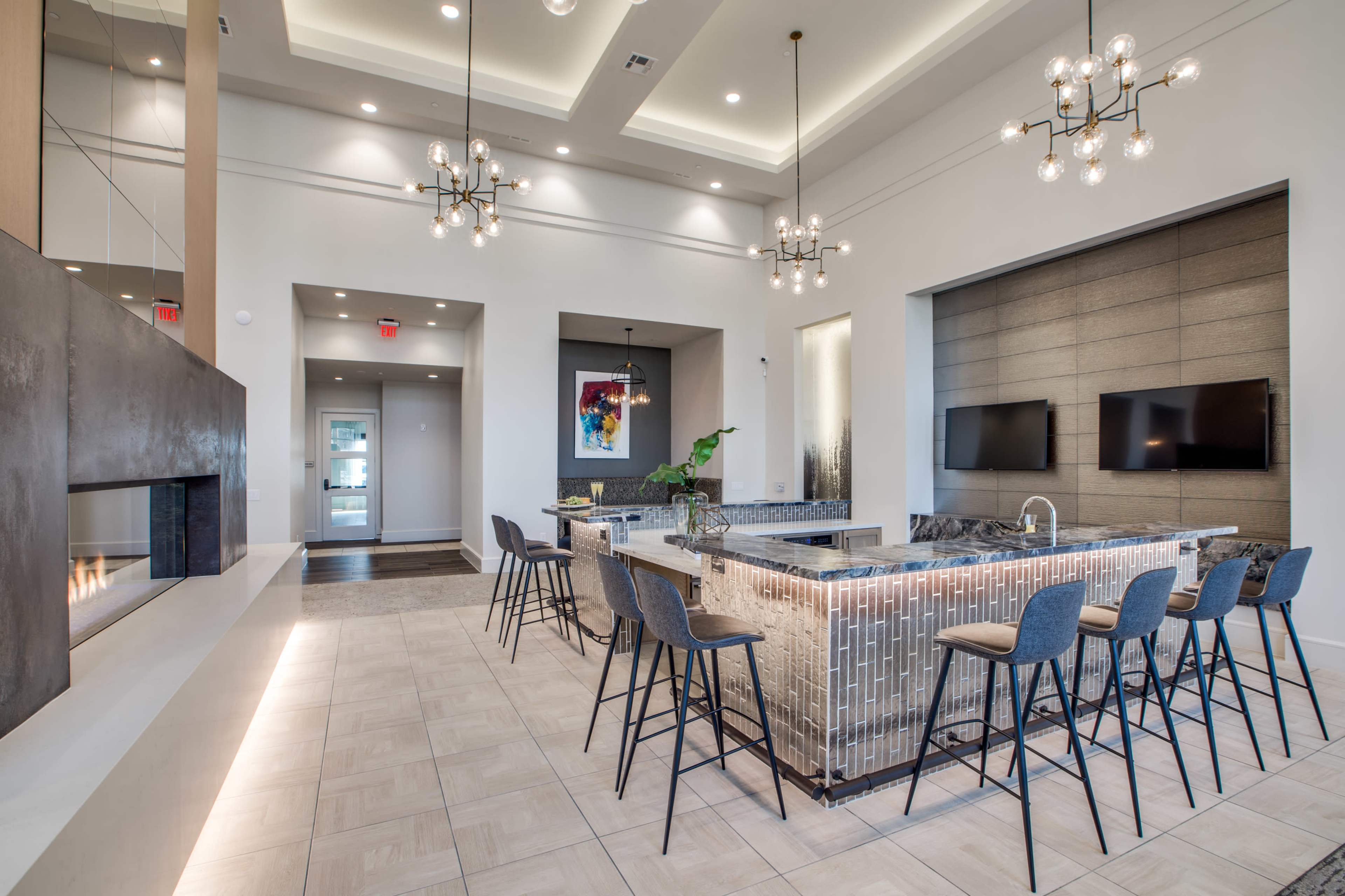 The image shows a modern lounge area featuring a kitchen island with barstool seating, a fireplace, and two wall-mounted televisions.