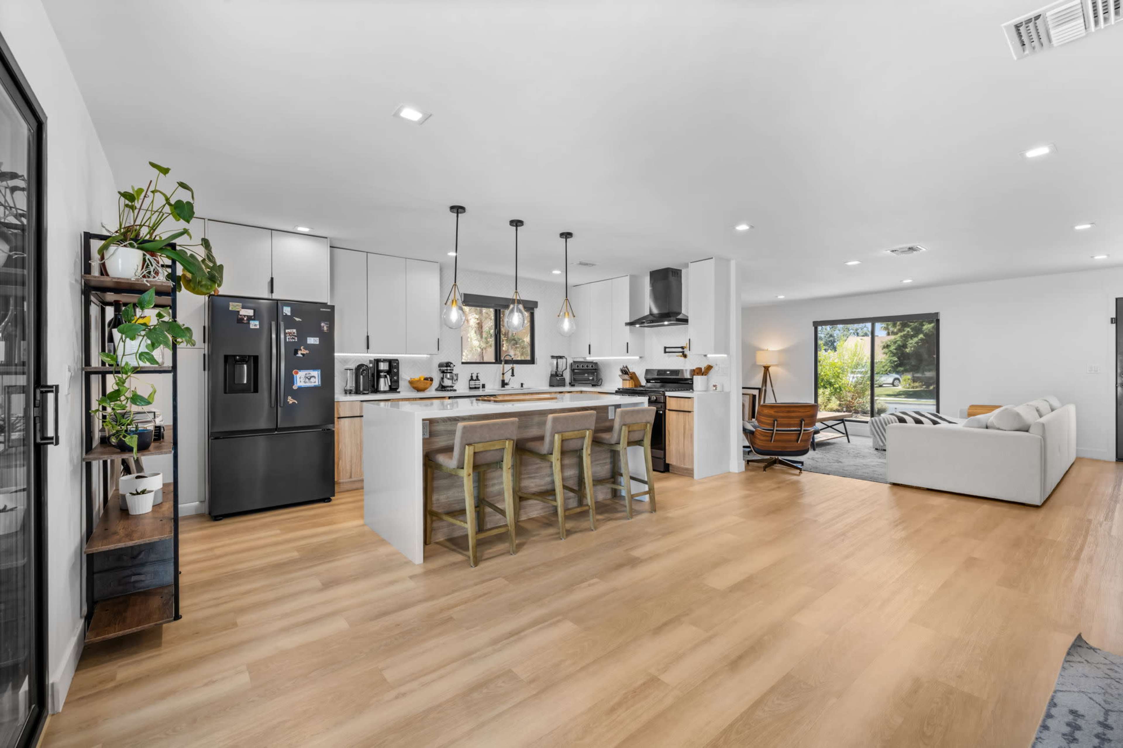 The image shows a modern open-concept kitchen and living area featuring a large island with seating, a black refrigerator, and a comfortable sofa facing a window.